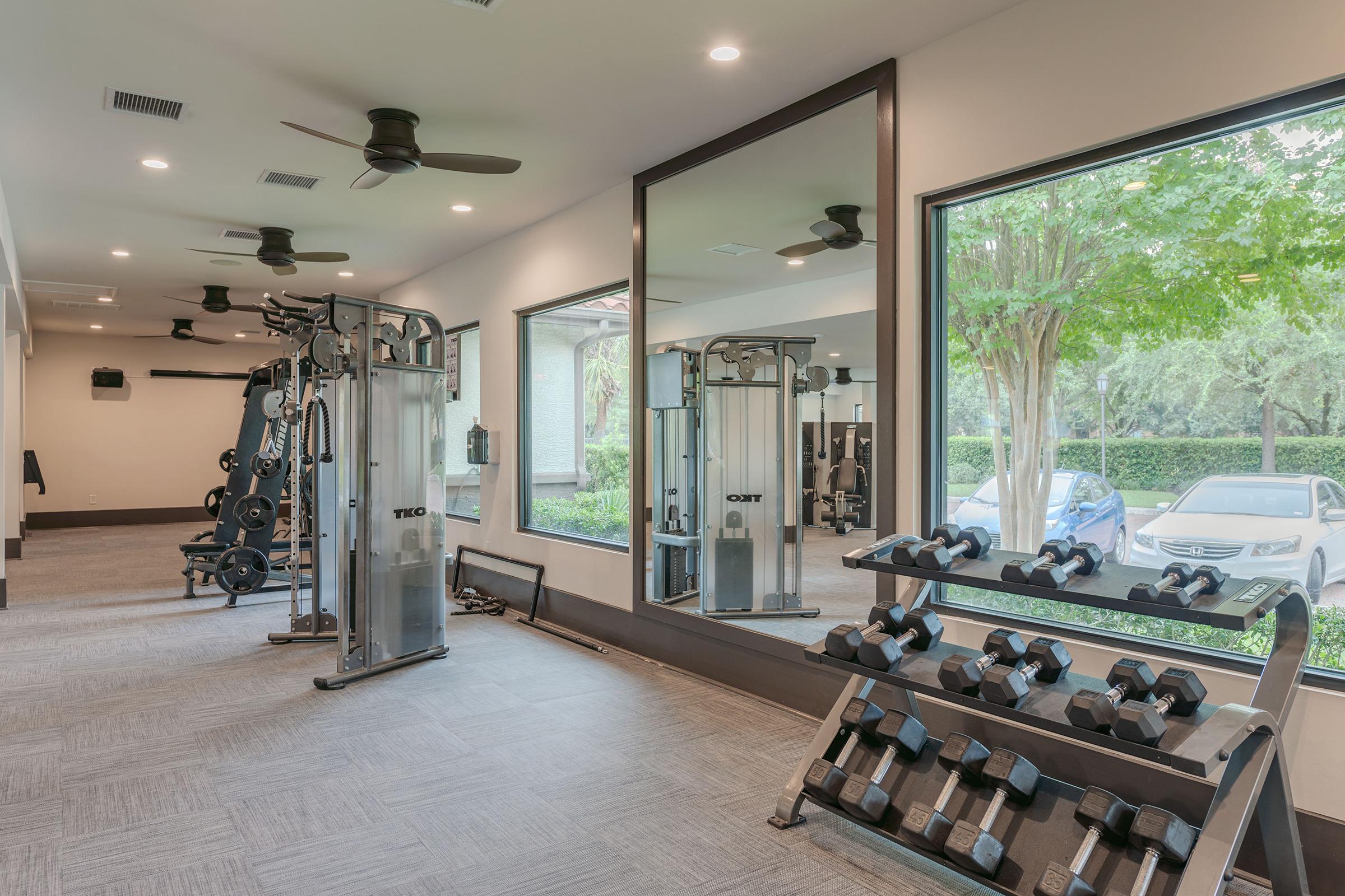 A modern gym interior featuring exercise equipment, including a weightlifting machine and a rack of dumbbells. Large windows provide natural light and a view of greenery outside. The space is well-lit and designed for fitness activities.