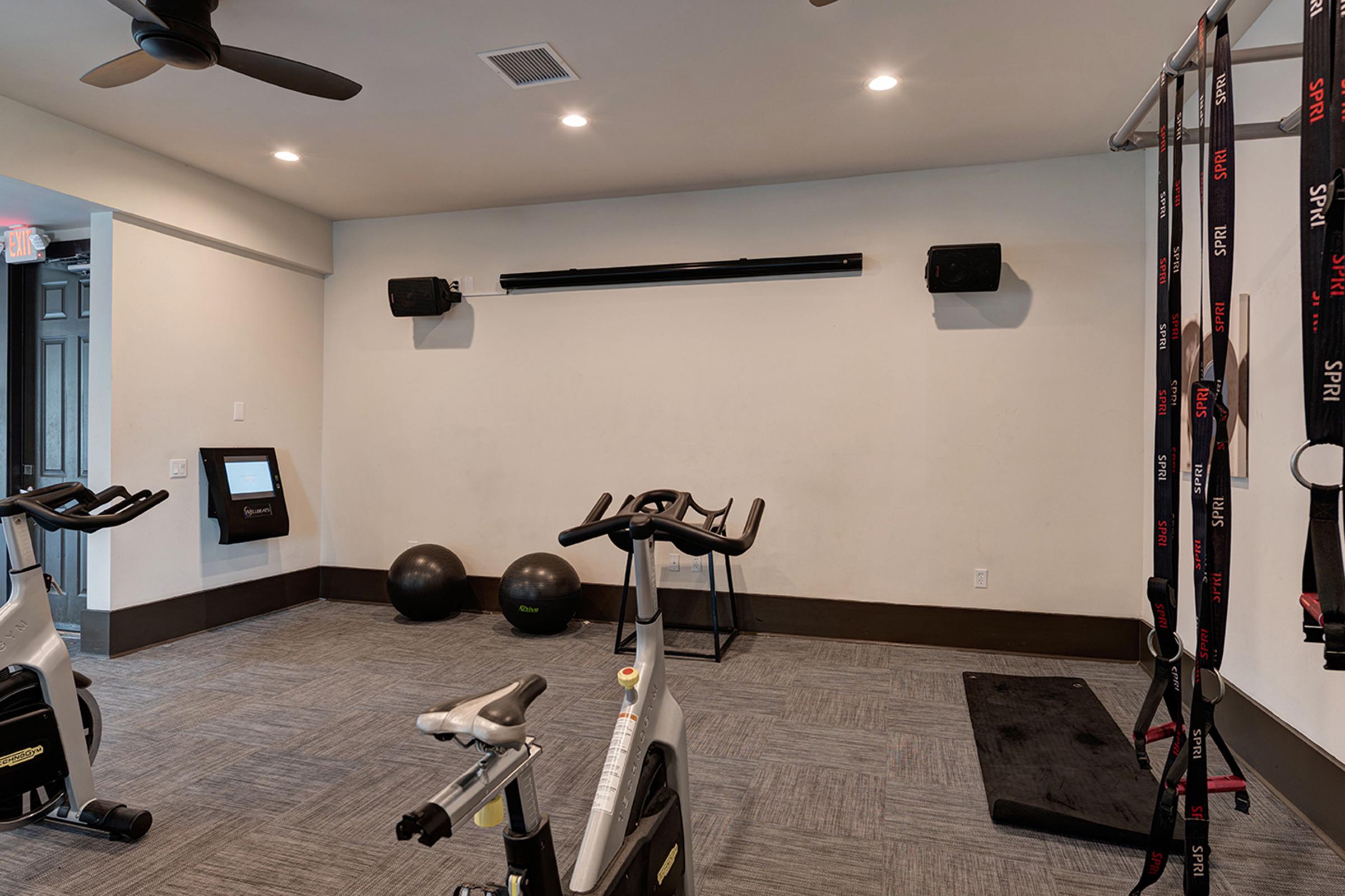 A well-lit exercise room featuring a stationary bike, two large exercise balls, a wall-mounted speaker system, and a wall with fitness straps. There's a touchscreen display on the wall, and the floor is covered with gray carpet tiles. The space has a clean, modern look with a minimalist design.
