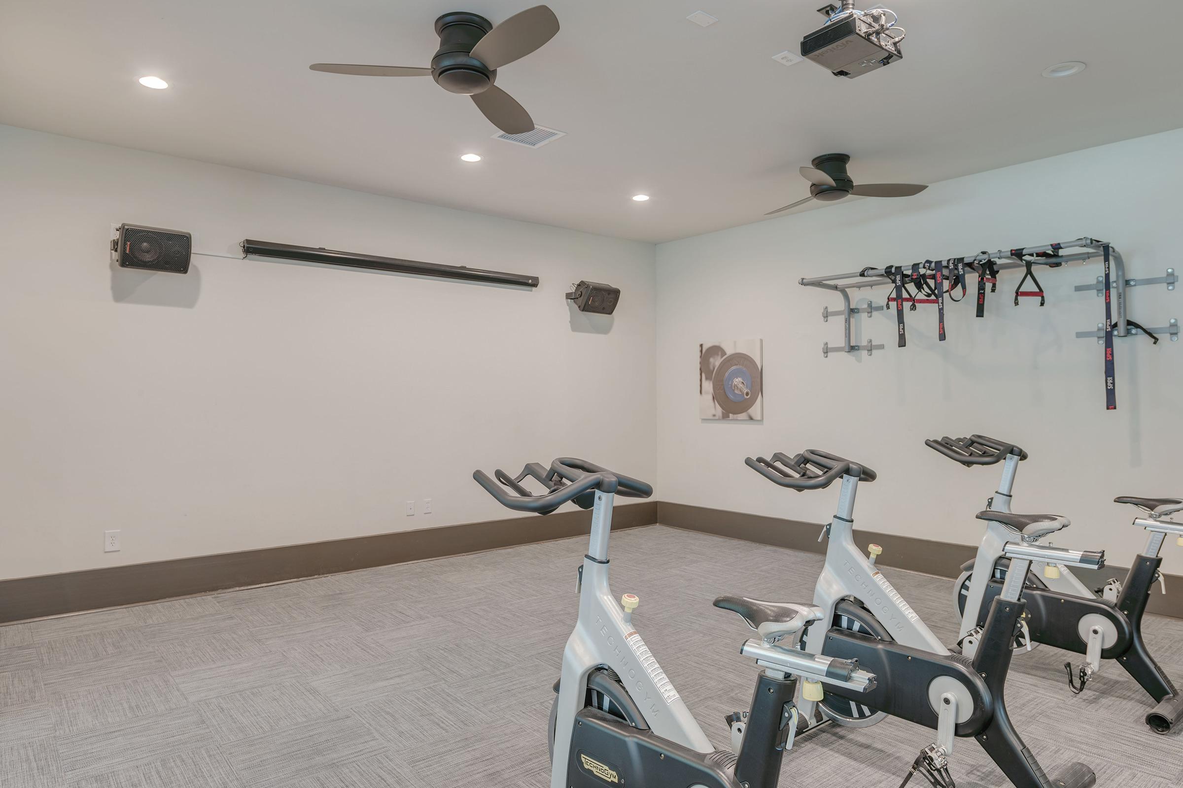 A modern fitness room featuring several stationary exercise bikes, a wall-mounted audio system, and suspension training equipment. The space has neutral-colored walls, ceiling fans, and a projector for workout guidance, creating an inviting environment for indoor cycling and fitness activities.