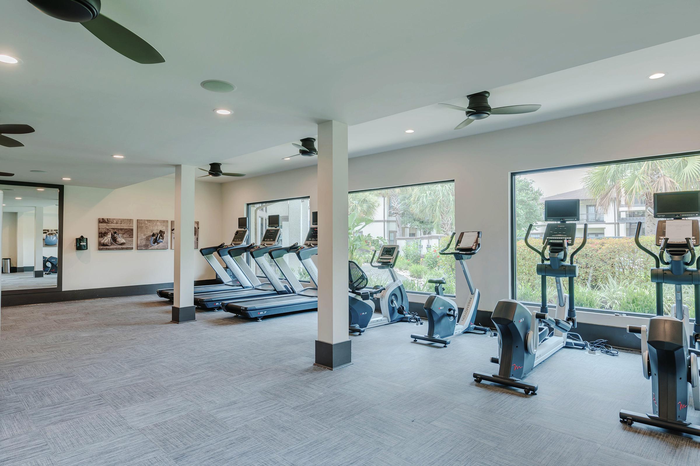 A modern fitness room featuring several treadmills and exercise bikes, large windows allowing natural light, and a view of greenery outside. The space is brightly lit and designed for a comfortable workout experience.