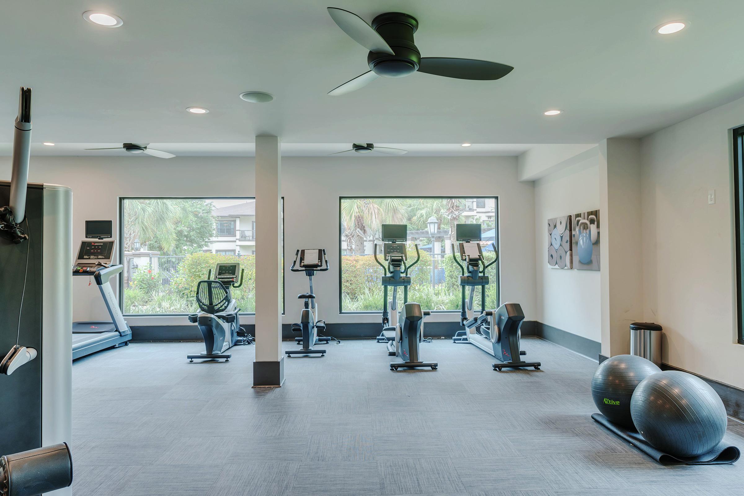 Interior of a modern gym featuring exercise equipment, including treadmills and stationary bikes. There are two large windows with views of greenery, and fitness balls placed on the floor. The space is well-lit with ceiling fans and a clean, contemporary design.