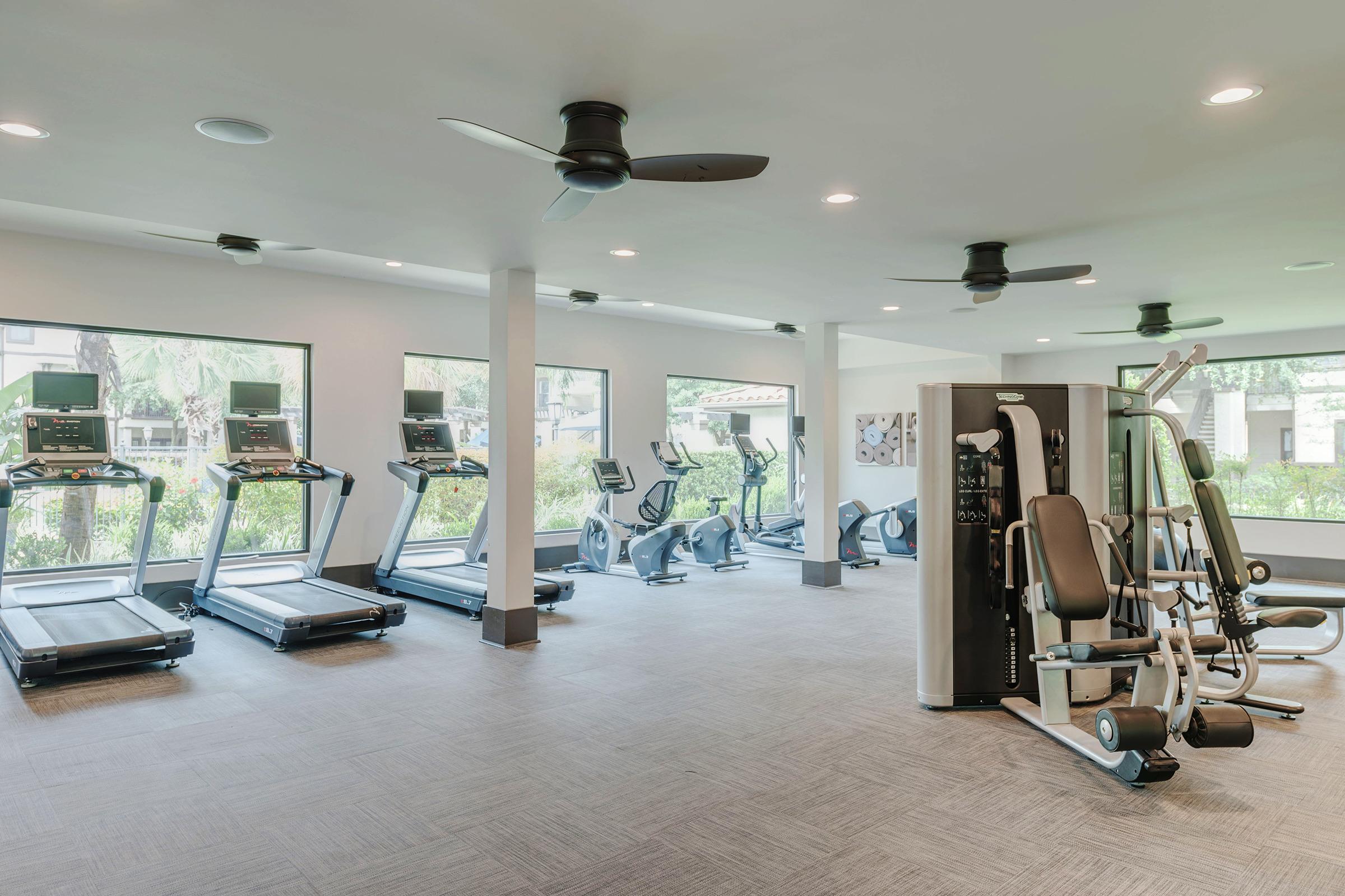 A modern gym interior featuring several treadmills, exercise bikes, and a strength training machine. The space is bright with large windows letting in natural light, equipped with ceiling fans for ventilation, and has a clean, minimalist design.