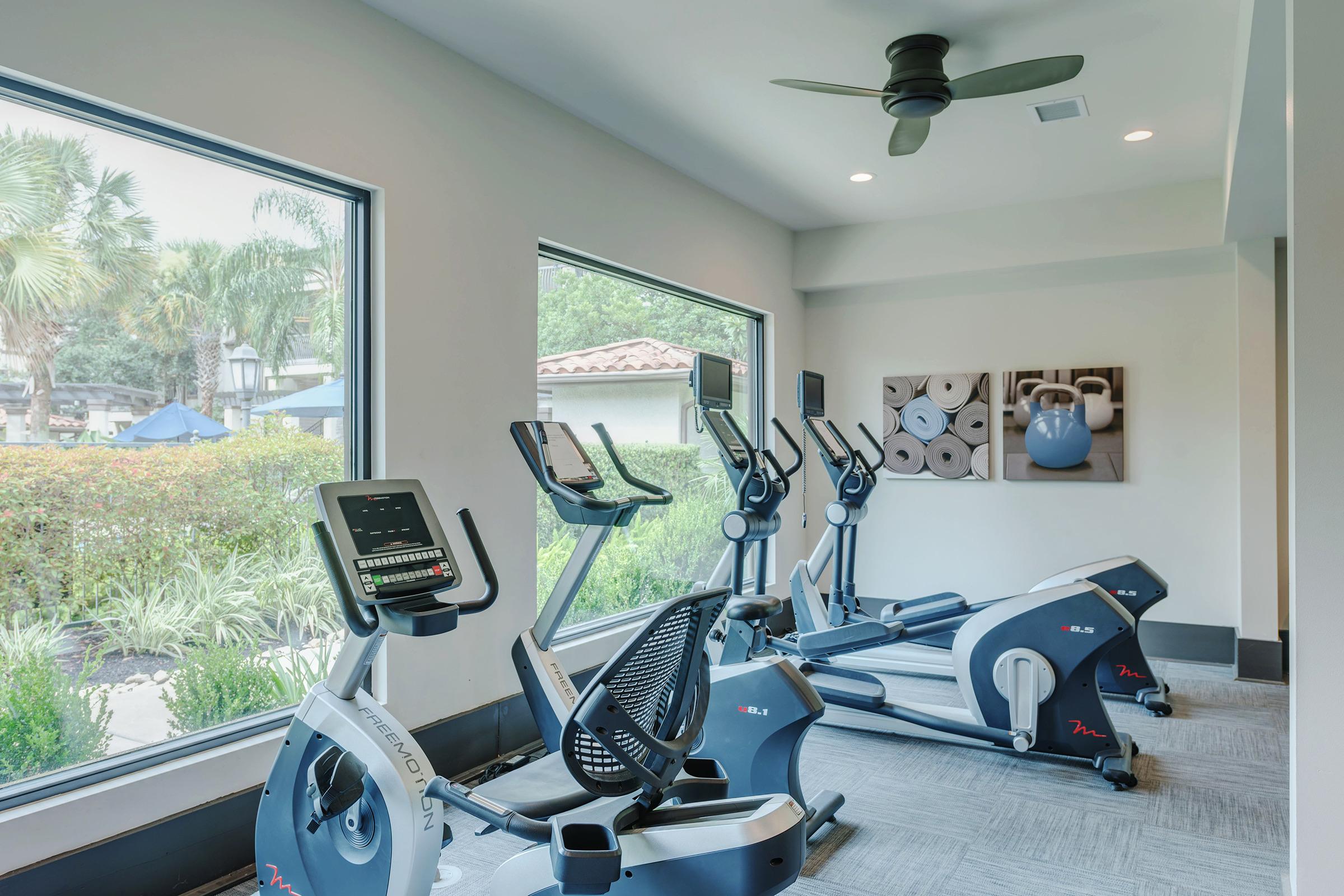A modern fitness room featuring several exercise machines, including stationary bikes and an elliptical trainer, with large windows overlooking greenery. A ceiling fan is visible, and there is a wall with framed blue kettlebells. The atmosphere is bright and inviting, ideal for workouts.