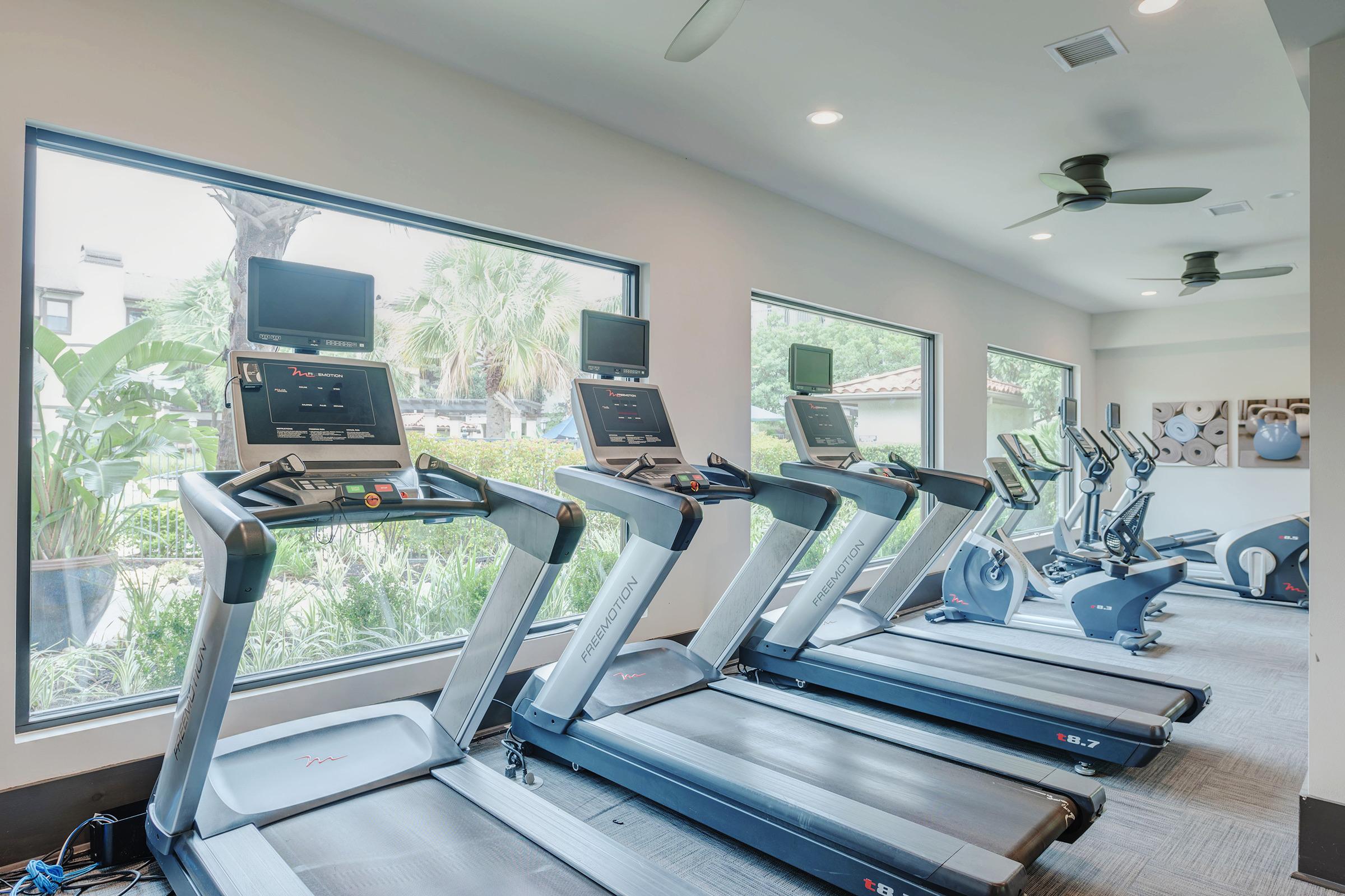 A modern gym interior featuring several treadmills and exercise bikes arranged near large windows, allowing natural light to fill the space. Green plants are visible outside, creating a refreshing atmosphere for workouts.