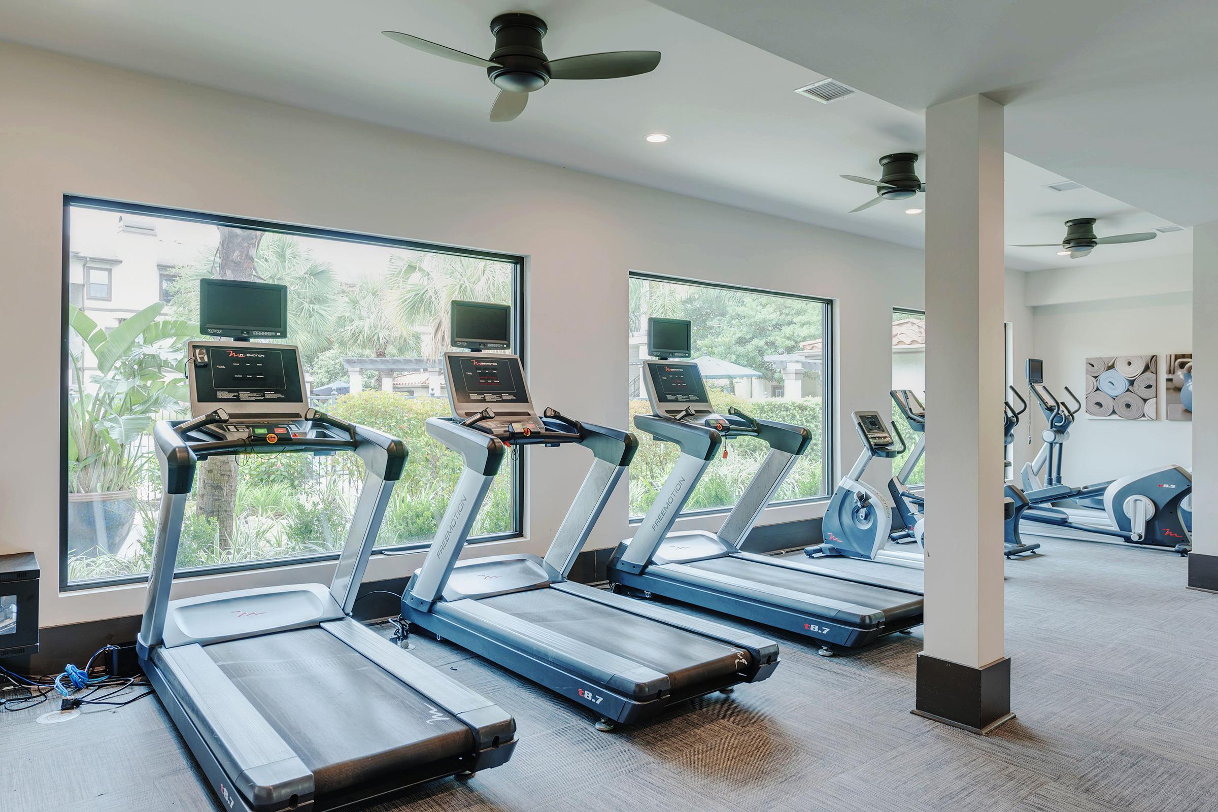 A well-lit gym area featuring four treadmills lined up against large windows, with greenery visible outside. The treadmills are modern, equipped with screens, and there are ceiling fans for ventilation. The space has a clean, contemporary design.