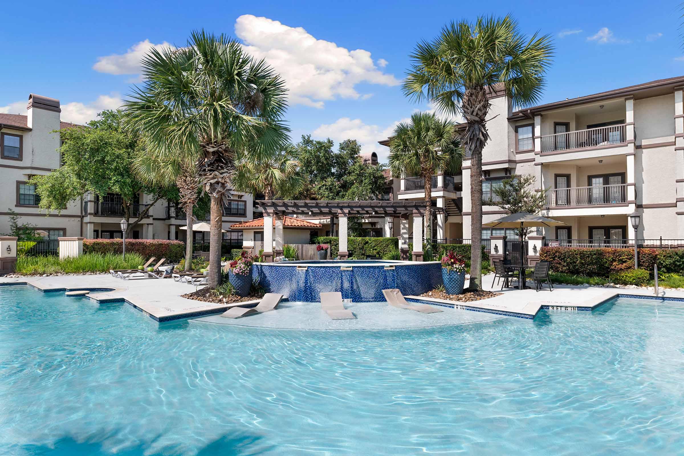 A bright, inviting pool area featuring crystal-clear water, surrounded by lush palm trees and landscaped greenery. In the background, there are modern apartment buildings with balconies. A spa-like seating area is centrally located in the pool, adorned with vibrant flowers. The sky is clear and blue, enhancing the tranquil atmosphere.