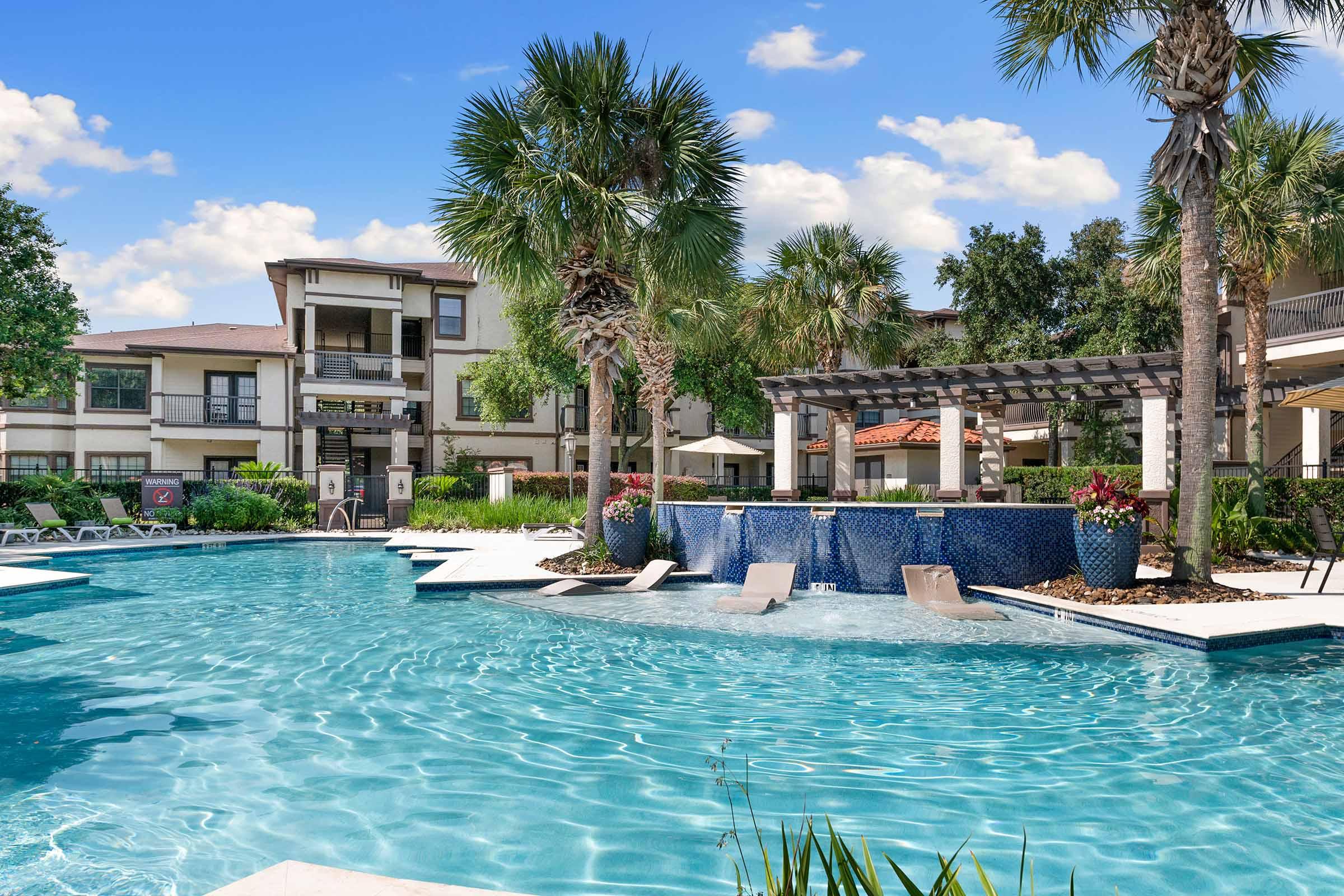A serene swimming pool area surrounded by lush palm trees and manicured landscaping. The clear blue water reflects the sky, while lounge chairs are placed neatly around the pool. In the background, an attractive residential building and a covered seating area add to the ambiance of relaxation.