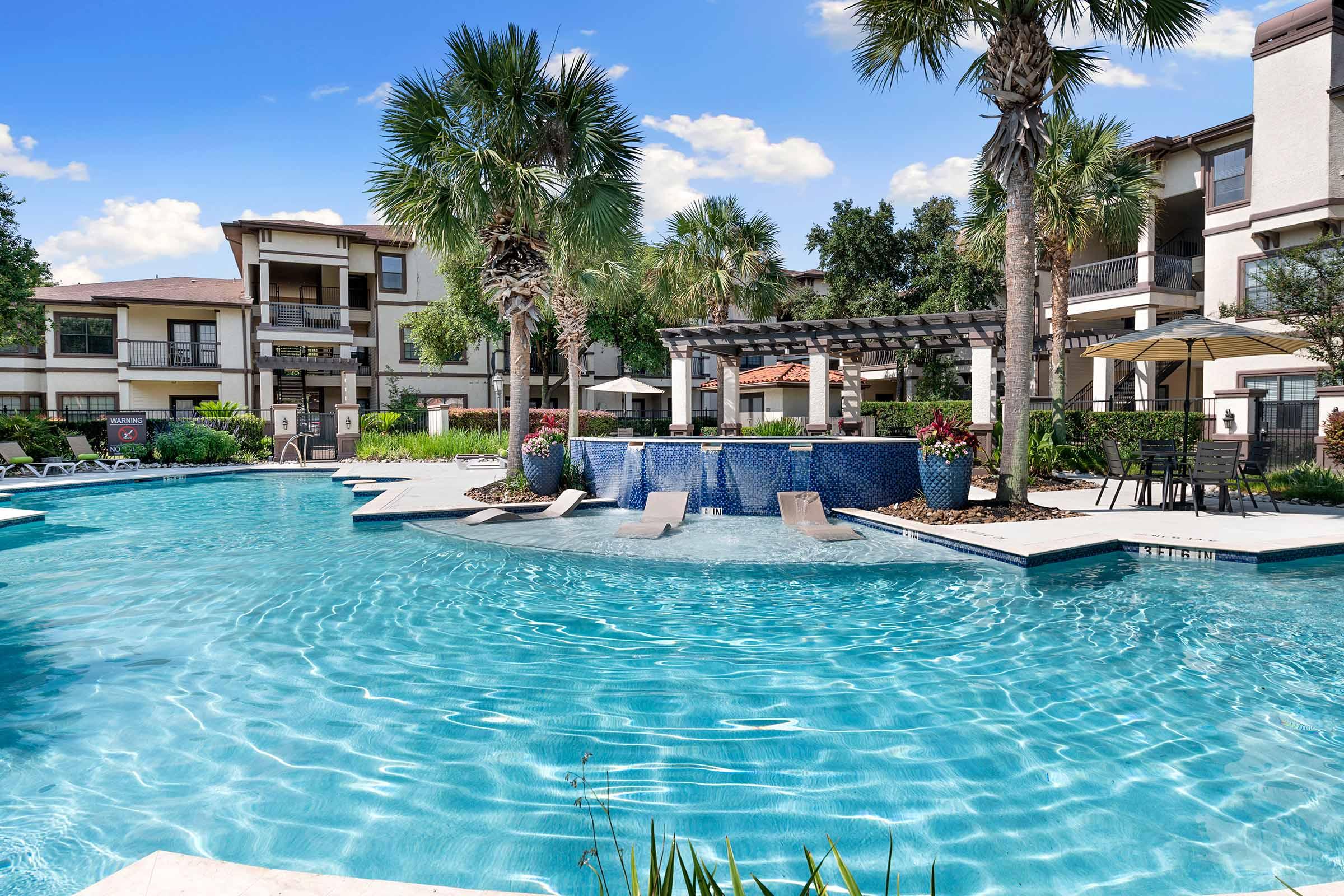 A vibrant swimming pool with clear blue water in a landscaped area featuring palm trees, lounge chairs, and a shaded seating area. Surrounding the pool are stylish apartment buildings, offering a relaxing outdoor space with lush greenery and a bright blue sky overhead.