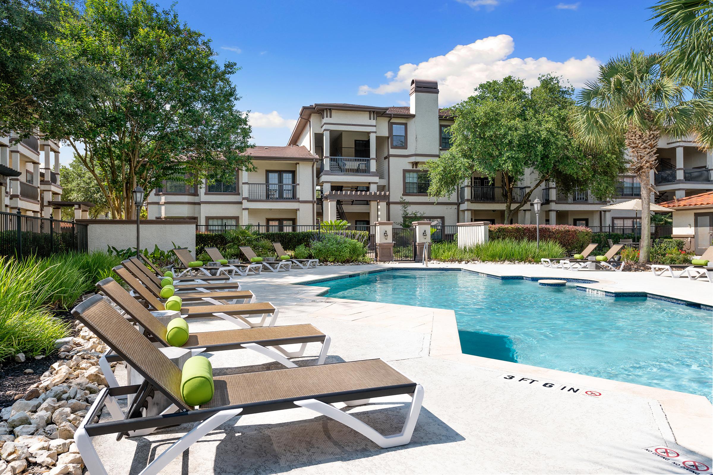 A serene outdoor pool area surrounded by lounge chairs and lush greenery. The pool features a shallow end marked with a depth sign, and the apartment complex is visible in the background, showcasing modern architecture against a clear blue sky.