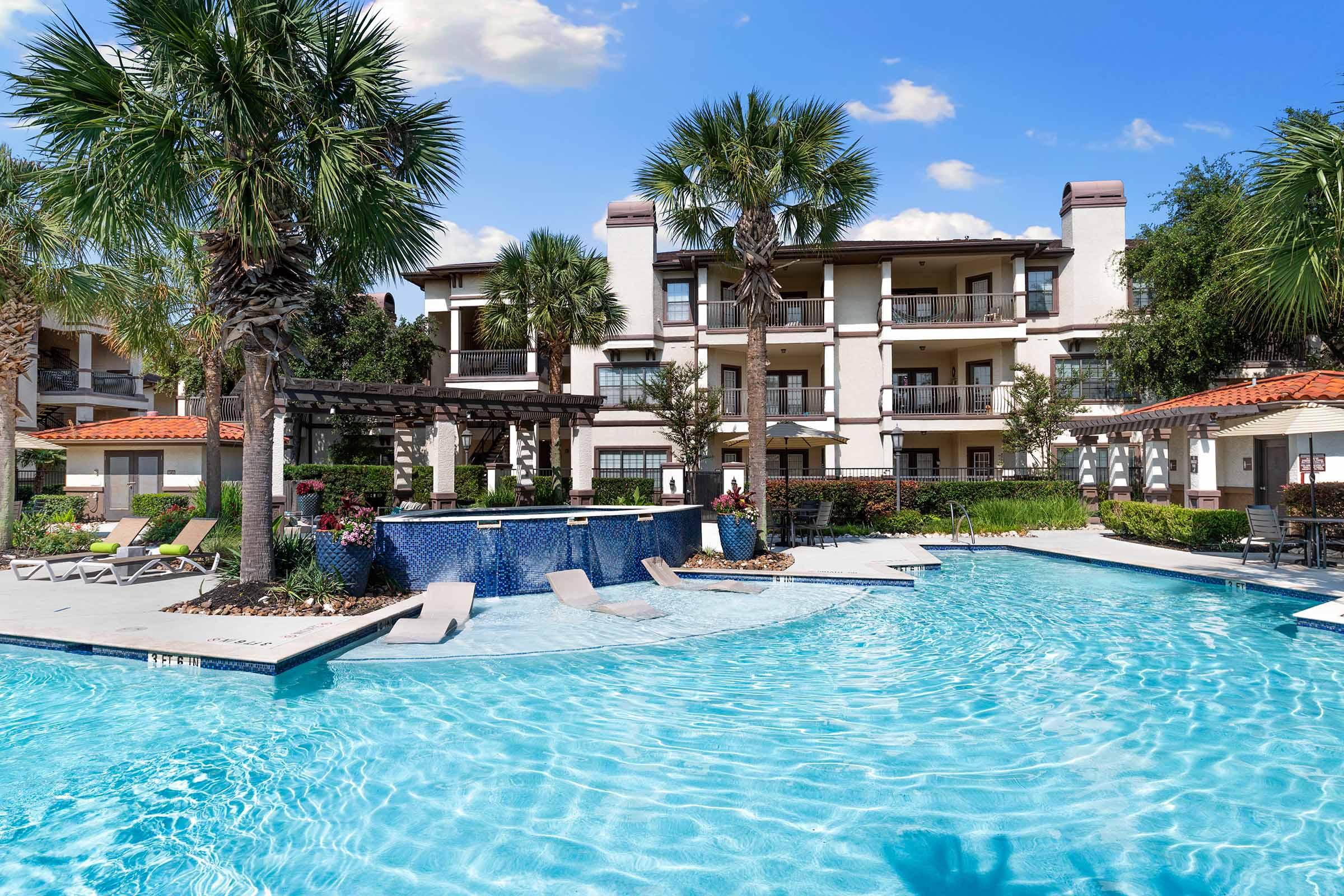 A serene swimming pool surrounded by palm trees, lounge chairs, and a spa area. In the background, a modern apartment building features balconies and well-maintained landscaping under a clear blue sky. The inviting pool is perfect for relaxation and leisure activities.