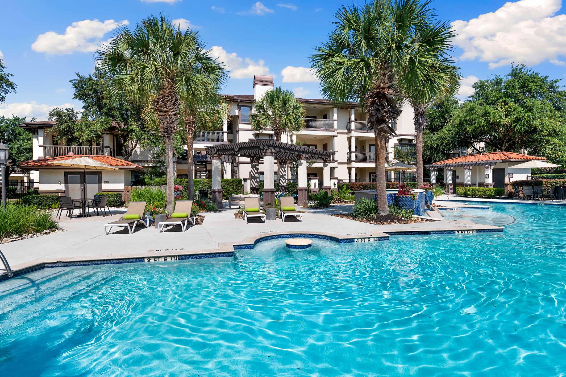 A vibrant outdoor pool area featuring crystal-clear water, surrounded by lounge chairs and palm trees. In the background, a multi-story apartment building with a pergola and greenery enhances the tropical ambiance, creating a relaxing atmosphere.
