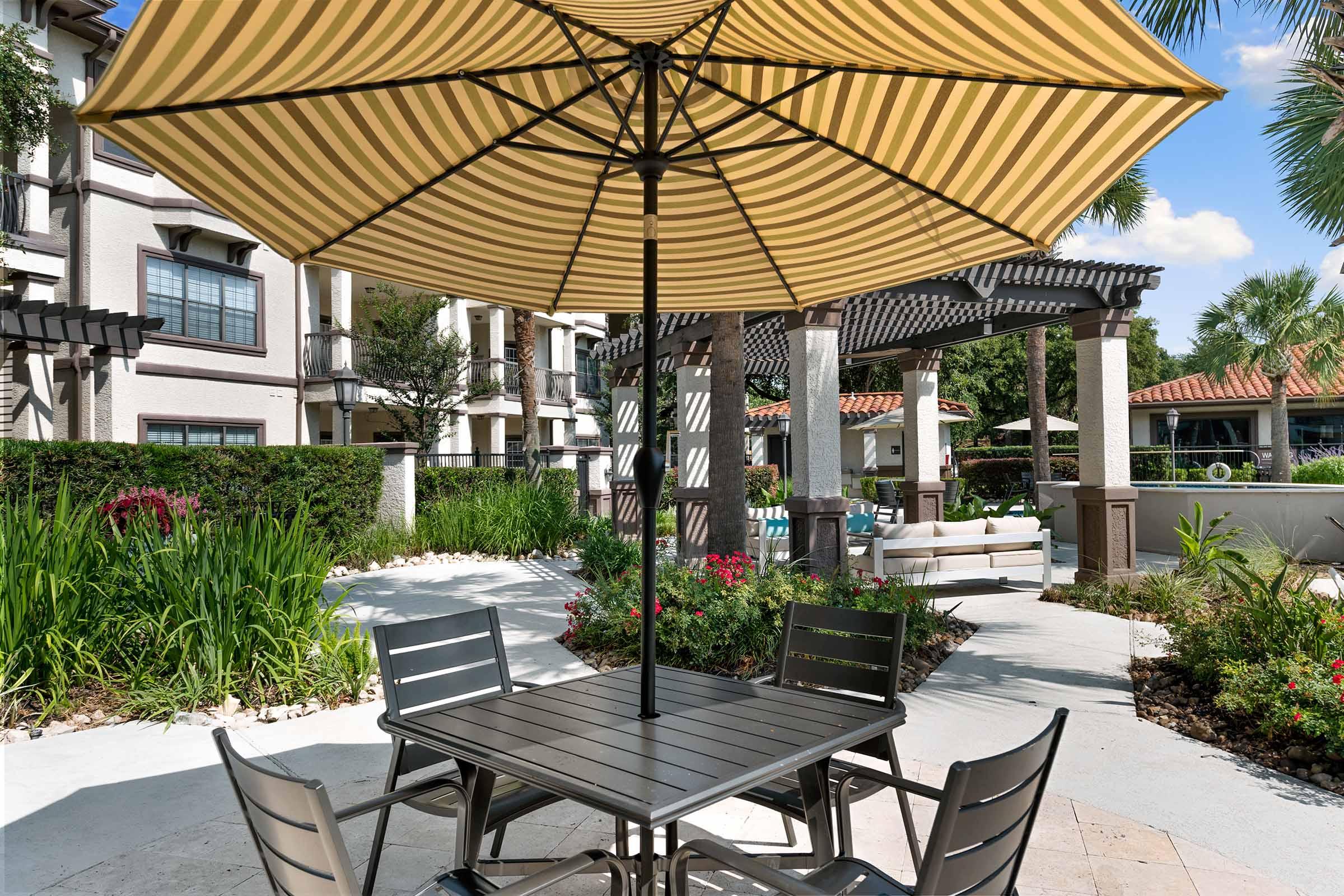 A shaded outdoor seating area featuring a striped patio umbrella, a black metal table, and chairs. The scene includes vibrant greenery, ornamental plants, and a background of residential buildings, creating a relaxed and inviting atmosphere.