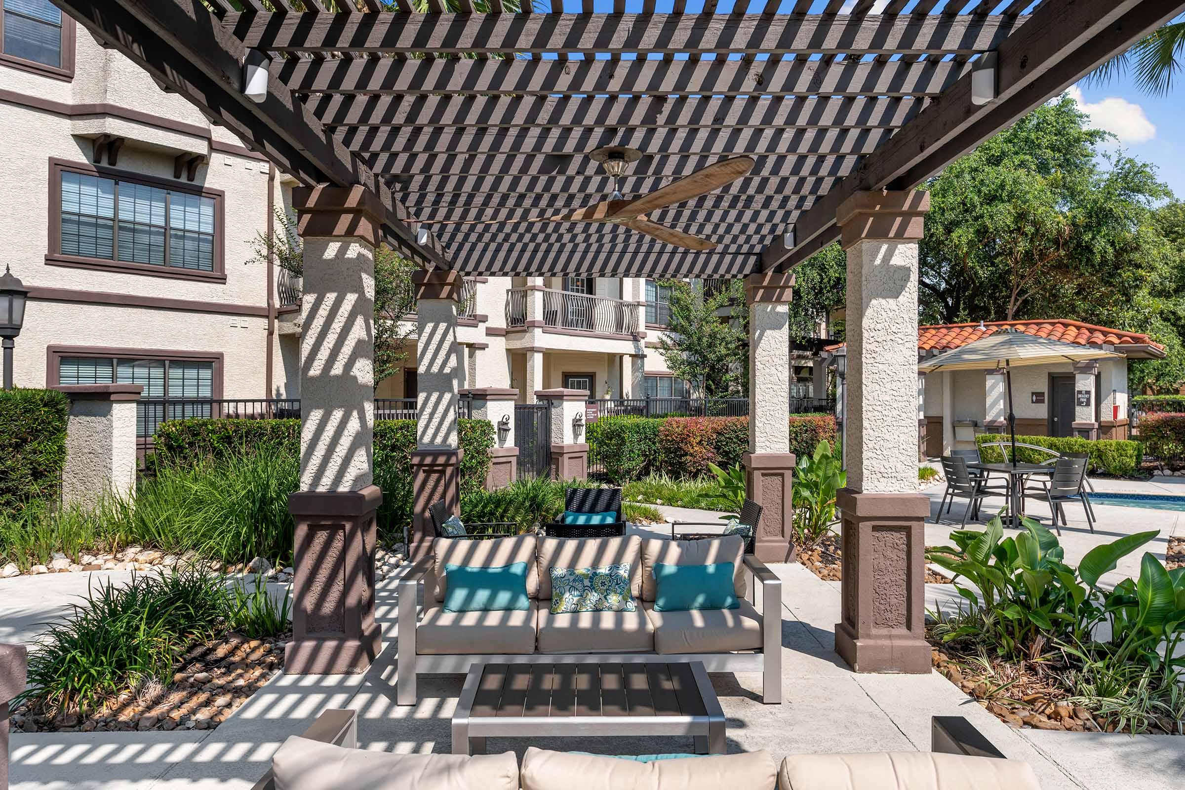 A shaded outdoor seating area featuring a modern furniture setup with cushions. Lush greenery surrounds the space, with a view of a nearby pool and a cabana in the background. Sunlight filters through a striped pergola, creating a relaxing atmosphere for leisure and socializing.