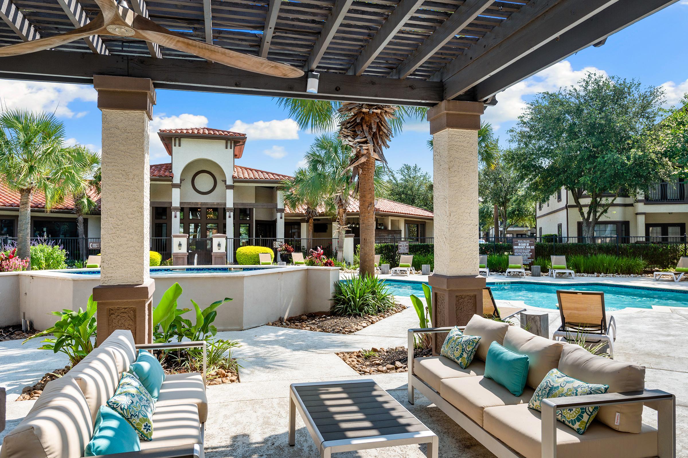 A serene outdoor lounge area featuring comfortable seating with colorful cushions, overlooking a sparkling swimming pool. Surrounding the pool are lush palm trees and well-maintained landscaping. In the background, there is a stylish clubhouse with a tiled roof, creating a relaxing atmosphere.
