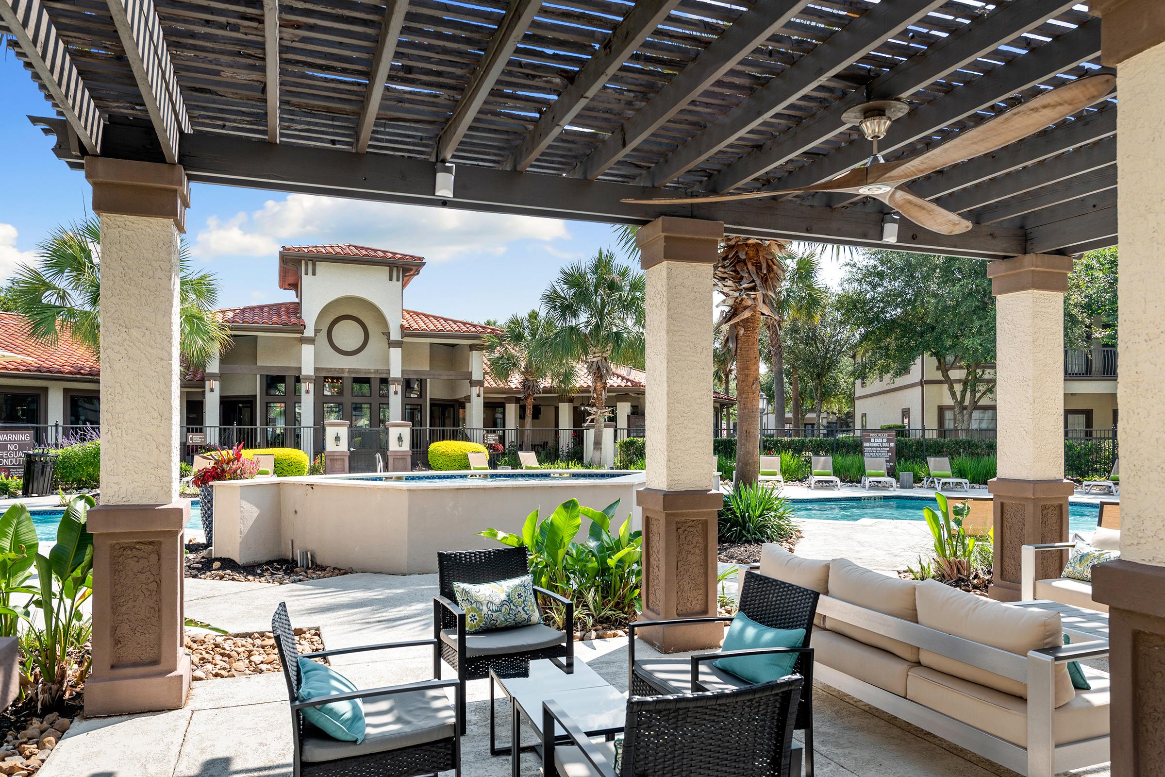 A sunny outdoor patio area with comfortable seating, featuring modern furniture, surrounded by lush greenery and palm trees. In the background, a well-maintained pool area and an elegant building with a red-tiled roof can be seen under a clear blue sky.