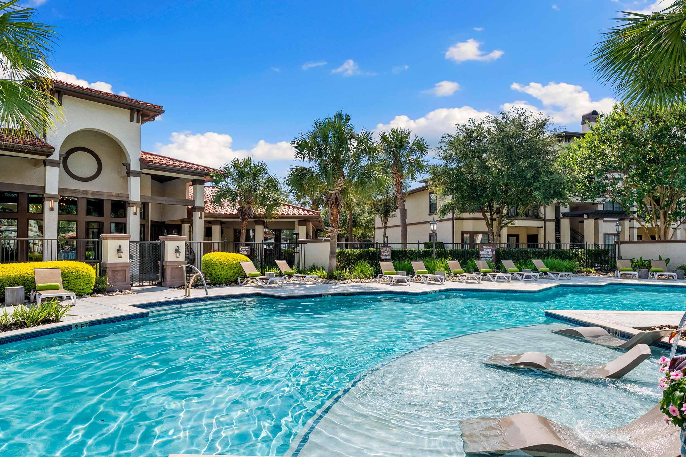 A sparkling blue swimming pool surrounded by lounge chairs, palm trees, and well-maintained landscaping. In the background, a stylish building with a Mediterranean design can be seen under a clear blue sky.