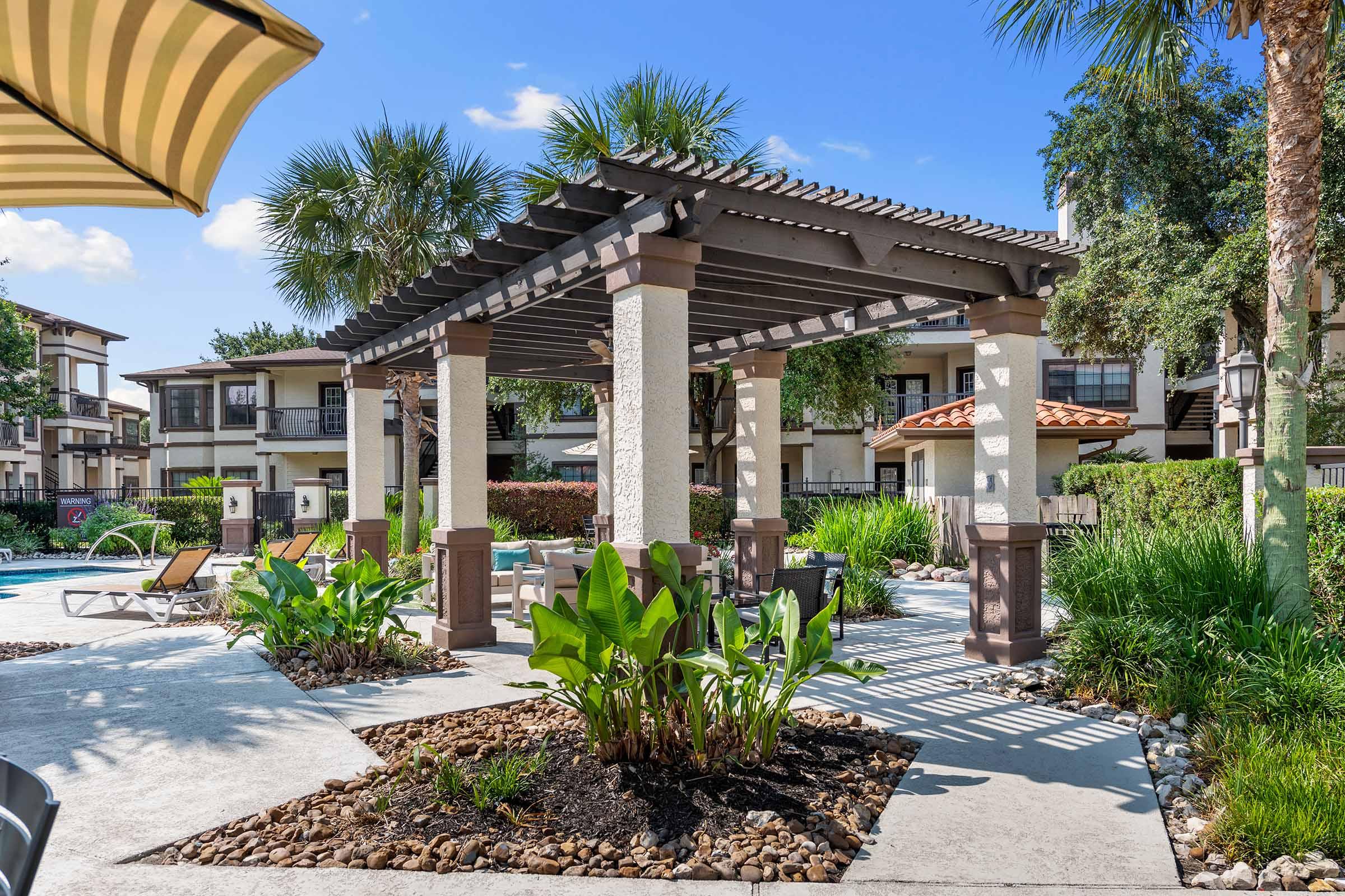 A landscaped pool area featuring a wooden pergola with seating, surrounded by palm trees and tropical plants. In the background, there are residential buildings with balconies, and the sky is clear and blue, creating a relaxing outdoor atmosphere.