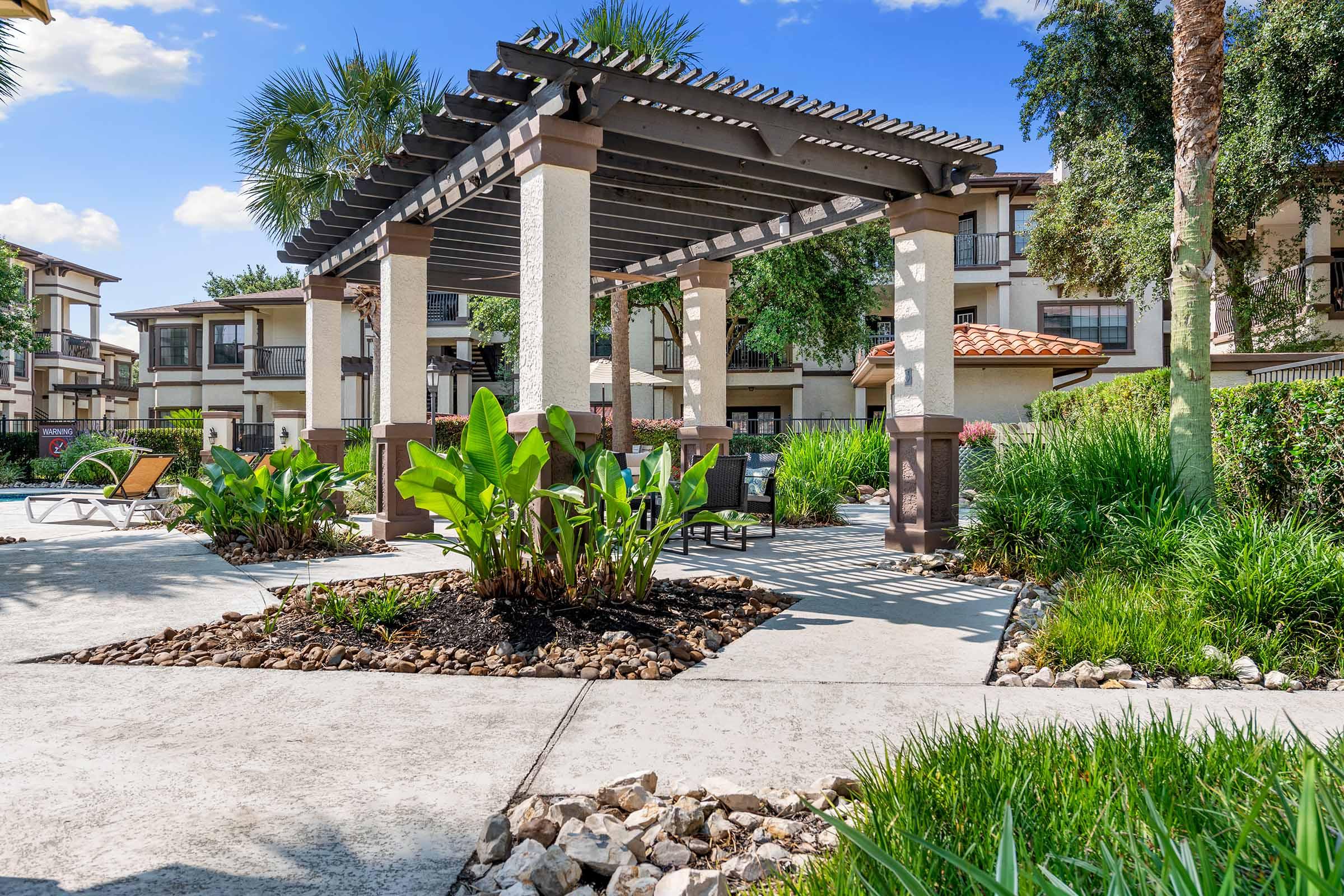 A landscaped courtyard featuring a wooden pergola surrounded by lush greenery, including palm trees and large leafy plants. Stone pathways and decorative rocks enhance the outdoor setting, with an apartment complex visible in the background under a clear blue sky.
