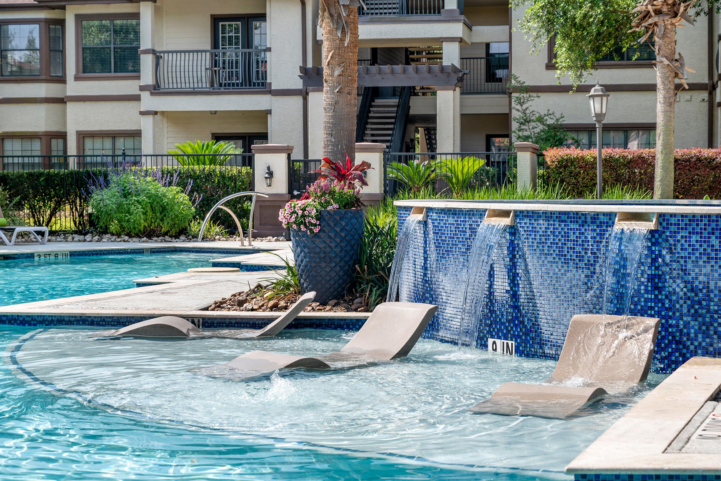 A serene swimming pool area featuring a blue mosaic tile wall with cascading waterfalls. There are several lounge chairs partially submerged in the water, surrounded by lush greenery and colorful plants. An apartment complex is visible in the background, providing a relaxing atmosphere.