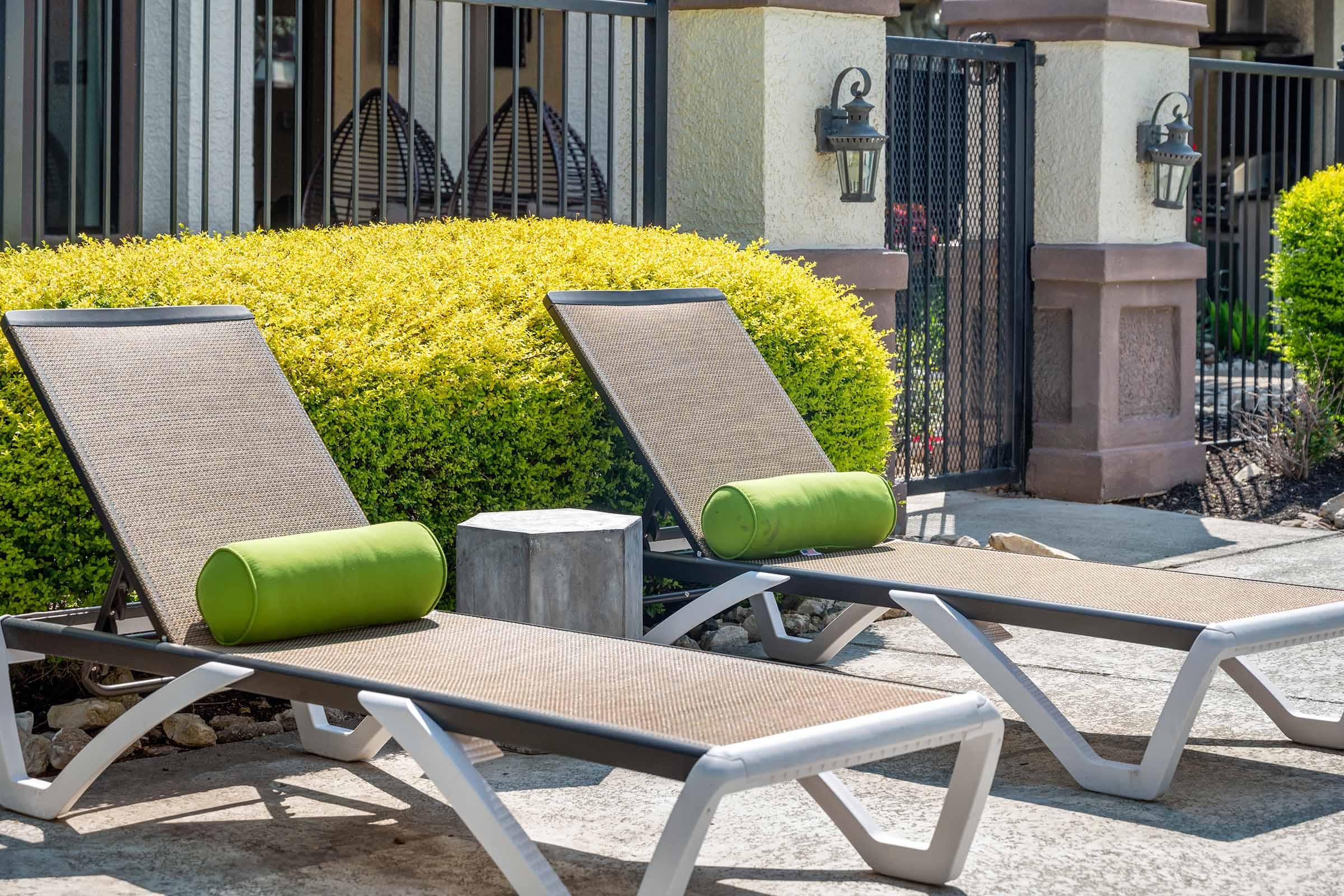 Two stylish lounge chairs with green cylindrical pillows are positioned beside a well-manicured hedge. The chairs are placed on a textured concrete surface, surrounded by a fenced area with decorative lanterns in the background. The scene conveys a relaxing outdoor space.