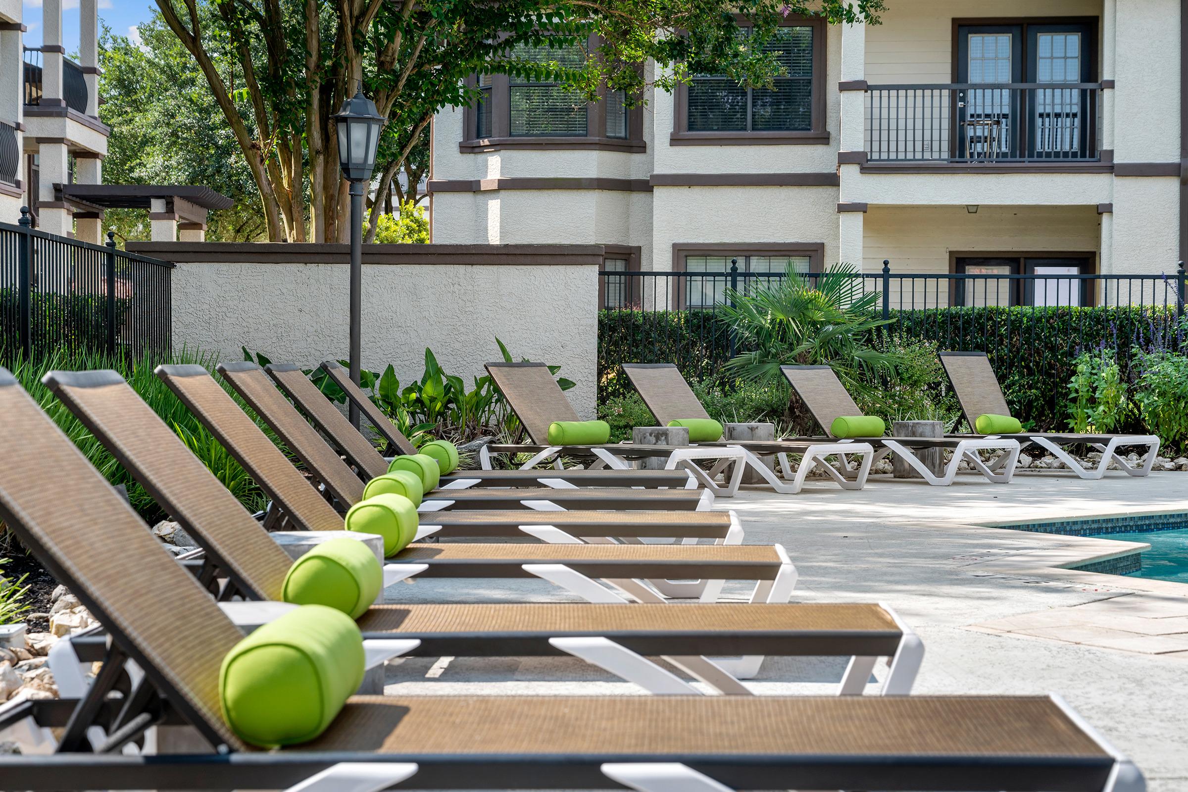 Loungers with green cushions lined up by a swimming pool, surrounded by lush greenery and a low fence. In the background, a multi-story building with balconies can be seen, creating a relaxing outdoor atmosphere. Sunlight brightens the scene, suggesting a warm and inviting pool area.