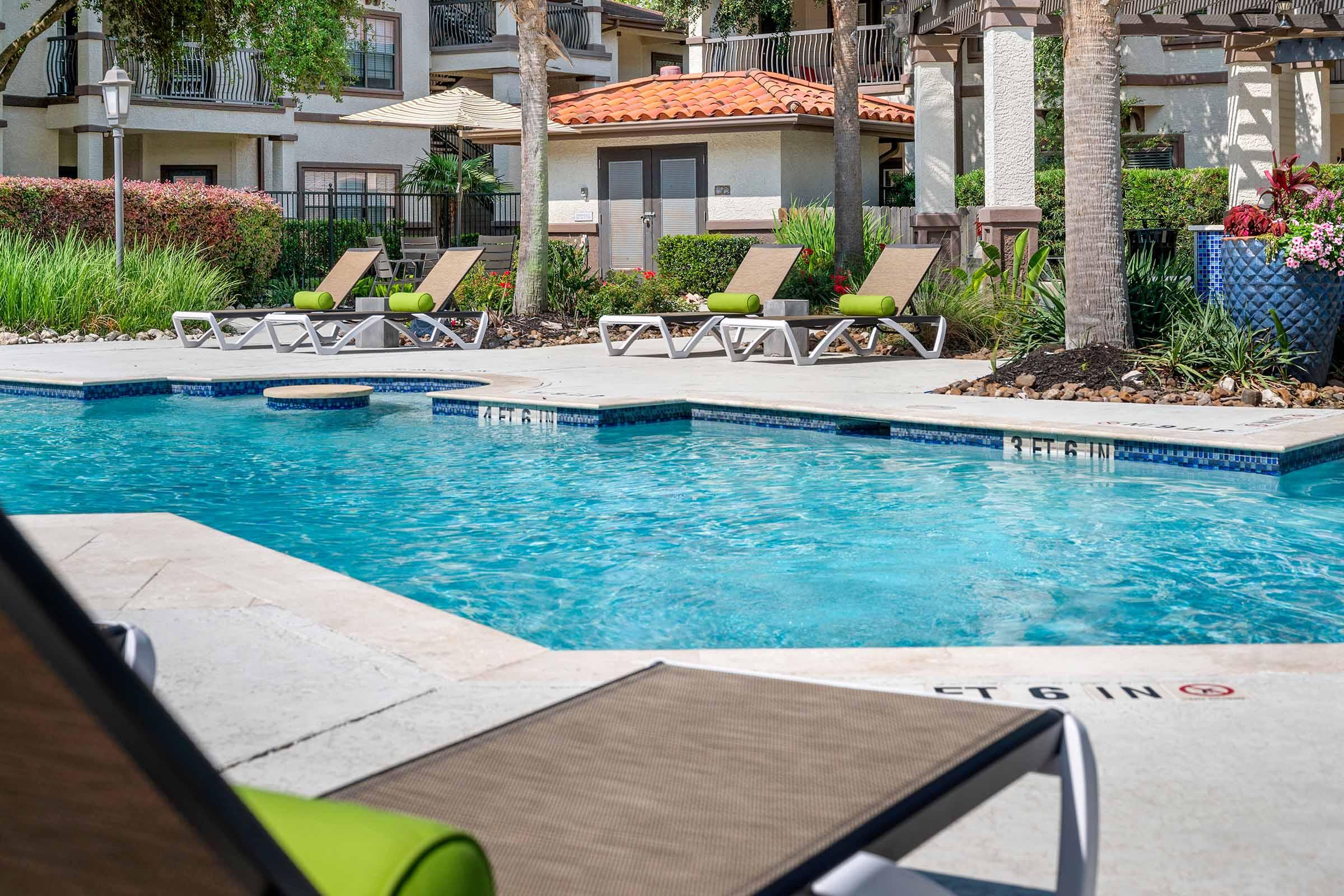A serene outdoor pool area featuring several lounge chairs, surrounded by lush greenery and palm trees. The clear blue water reflects sunlight, while nearby buildings and a patio area create a relaxing atmosphere.