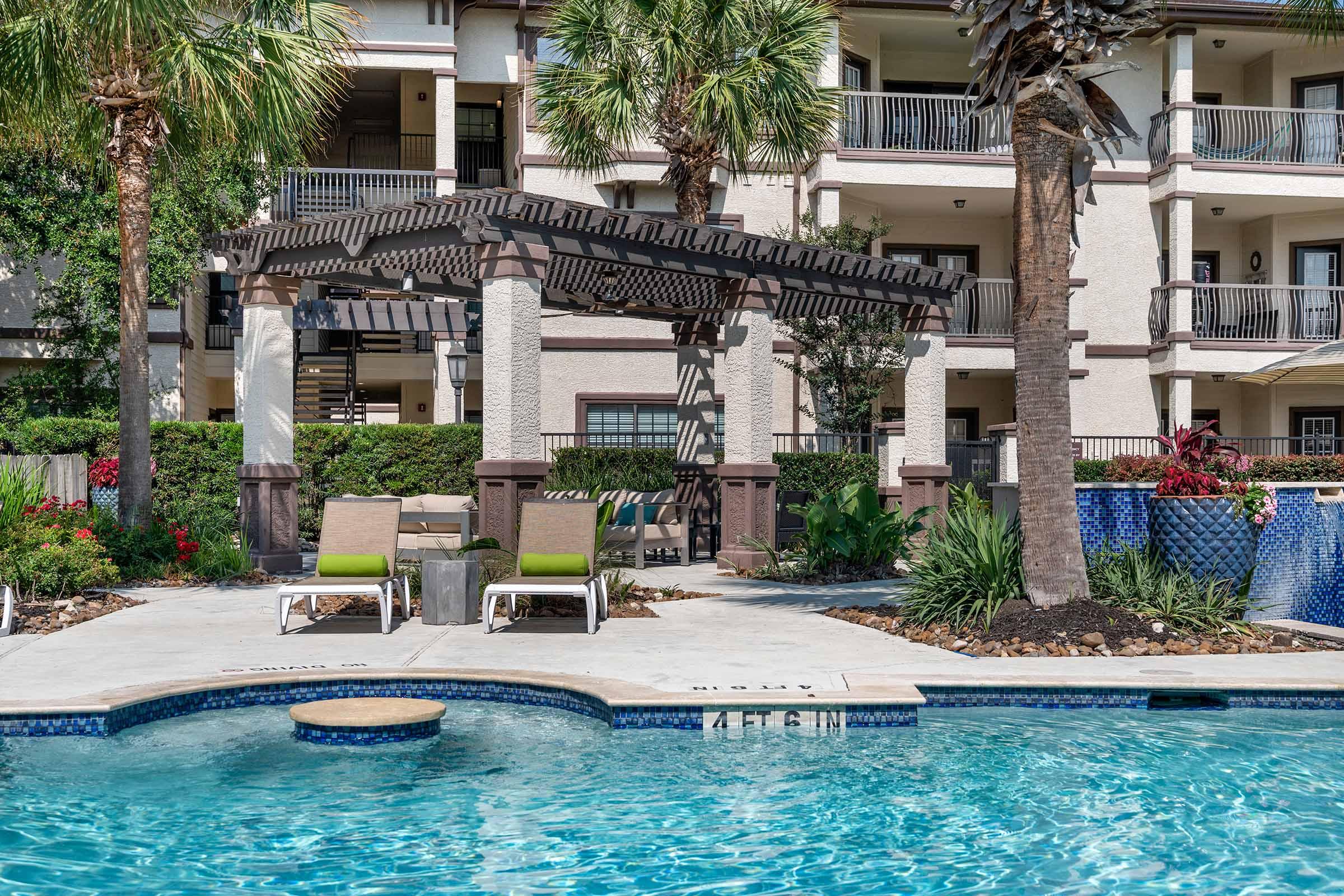 A swimming pool surrounded by tropical landscaping and lounge chairs. In the background, there is a shaded pergola with seating. The pool features a circular area for seating, and there are palm trees and colorful flowers enhancing the outdoor space of an apartment complex.
