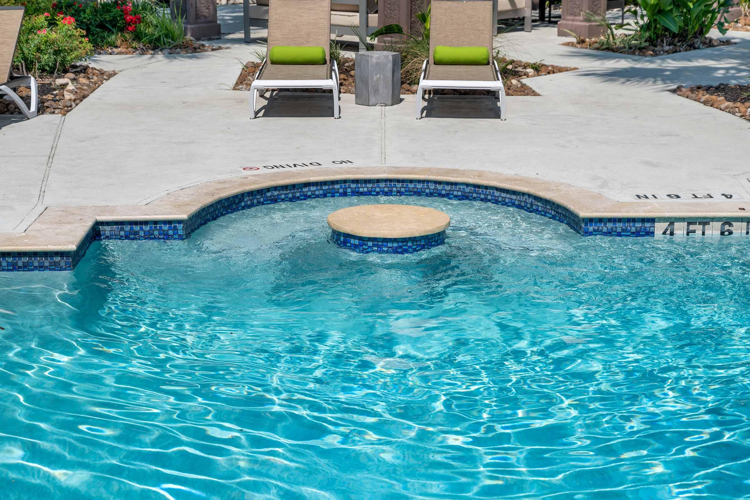 A clear blue swimming pool with a circular platform in the center, surrounded by light-colored concrete. Lounge chairs with green cushions are positioned nearby, and vibrant flowers and greenery are visible in the background. Sunlight reflects off the water, creating a sparkling effect.