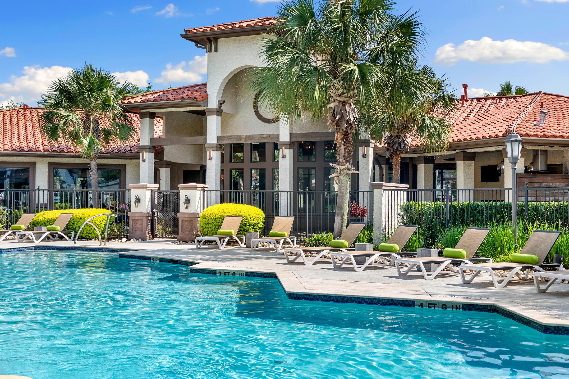 A sunny pool area featuring turquoise water surrounded by lounge chairs. In the background, a modern building with a red tile roof and palm trees adds a tropical vibe. Lush greenery and landscaped bushes enhance the inviting atmosphere, perfect for relaxation.