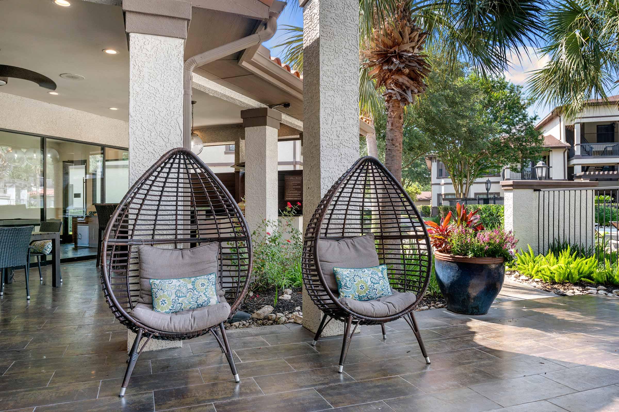 Two modern, teardrop-shaped wicker chairs with gray cushions and decorative pillows are placed outdoors. They sit on a tiled patio surrounded by lush greenery and palm trees, with a glimpse of a building in the background, creating a welcoming and stylish outdoor seating area.