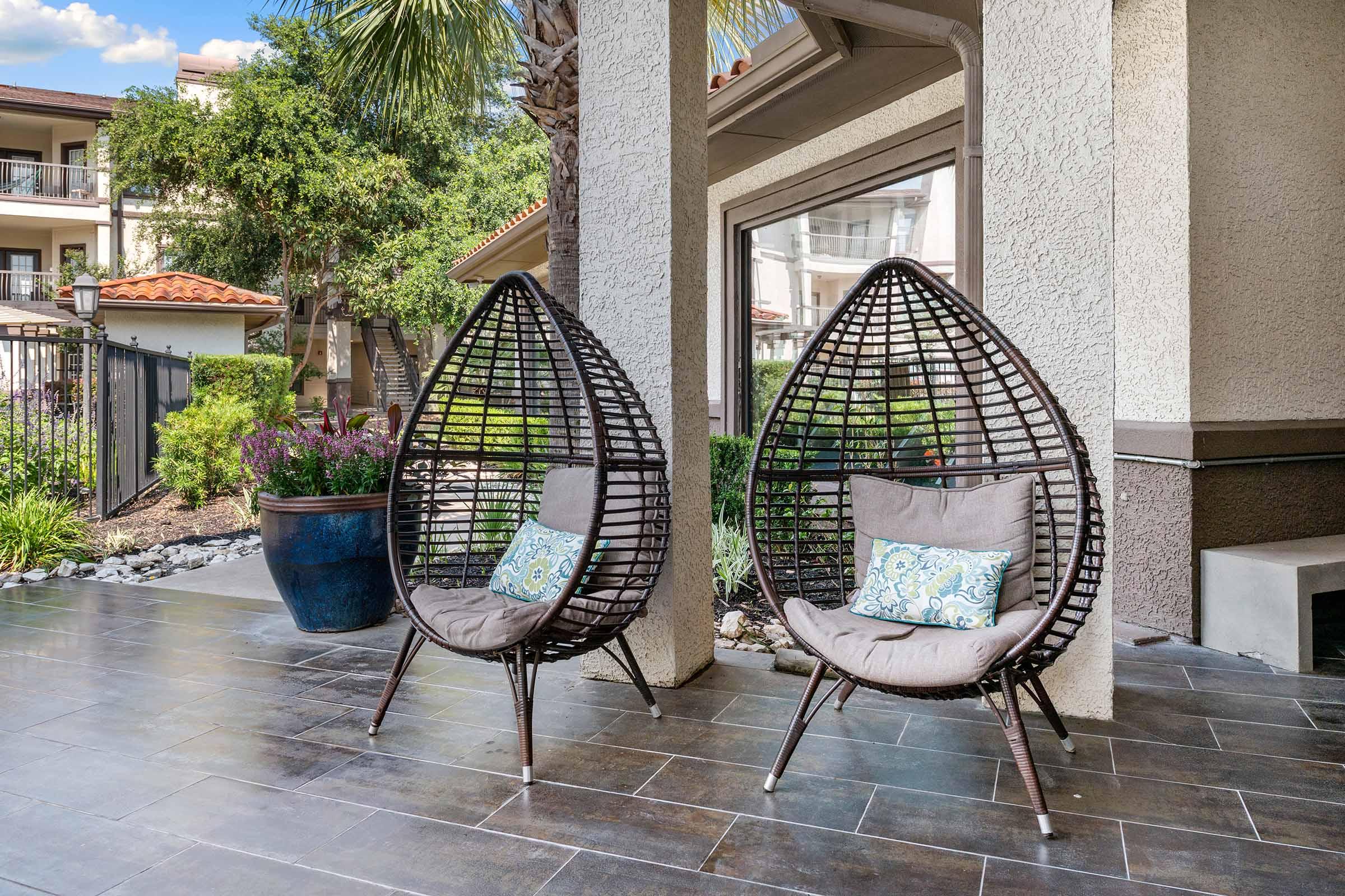 Two modern, teardrop-shaped chairs made of dark wicker are positioned on a tiled patio. Each chair features a soft cushion with a blue and white decorative pillow. In the background, lush greenery and a building can be seen, creating a serene outdoor space.