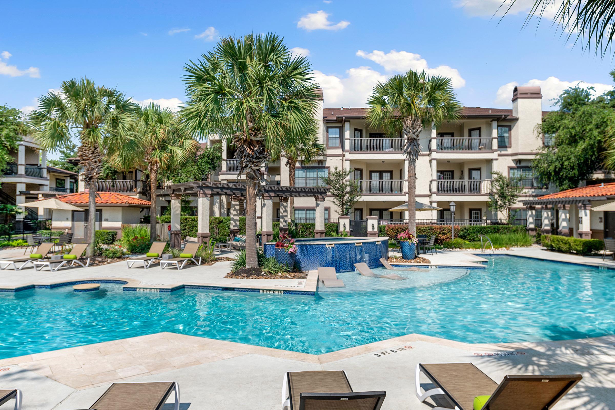A sunny outdoor pool area surrounded by palm trees and lounge chairs, with a fountain and lush landscaping. The pool is located in front of a multi-story apartment building with balconies, creating a relaxing and inviting atmosphere. Blue skies and white clouds add to the pleasant ambiance.