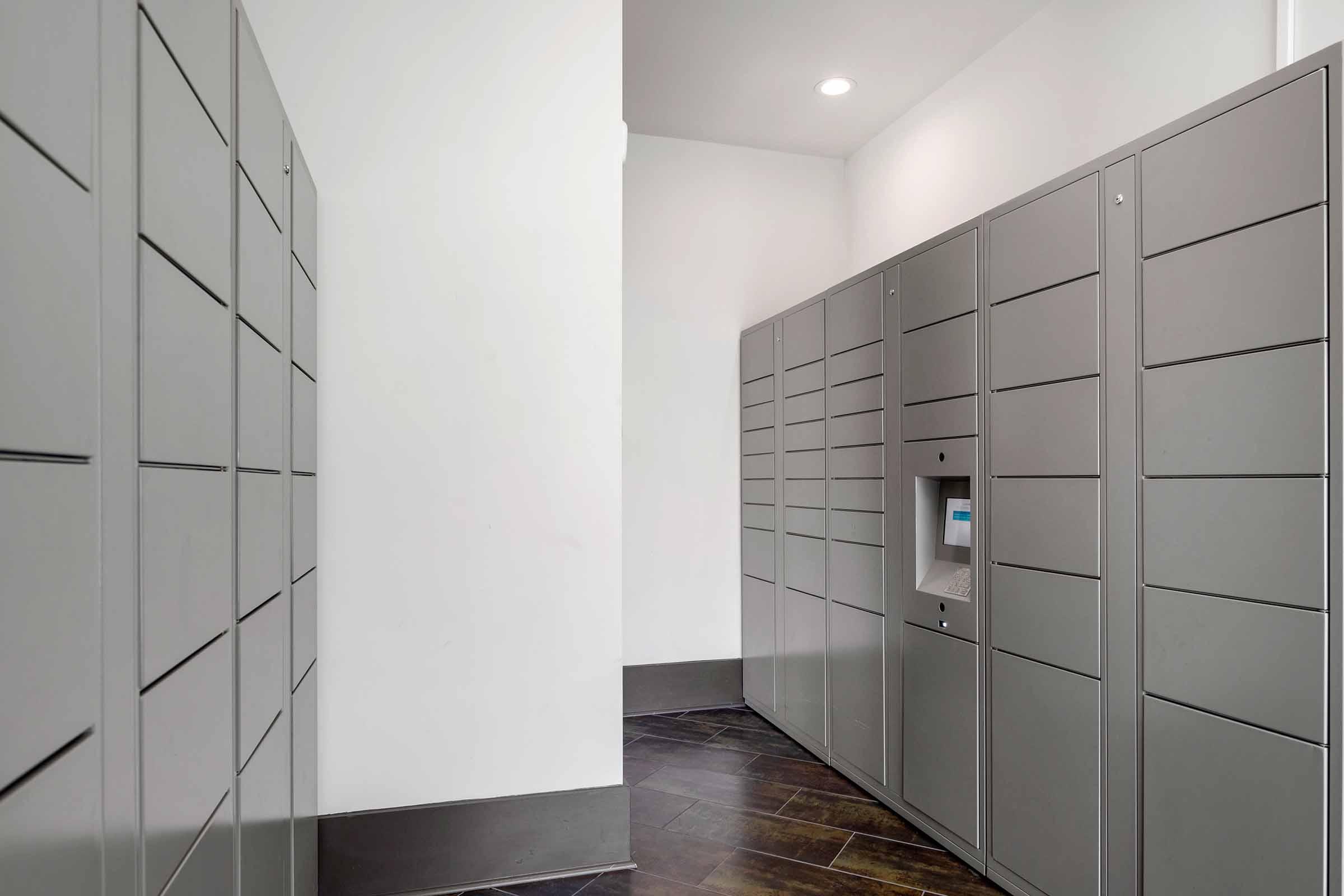 A modern hallway with gray parcel lockers lining the walls. The lockers are uniform in design and arranged in rows, with a digital interface on one of the lockers. The space is well-lit and has a clean, minimalist aesthetic.
