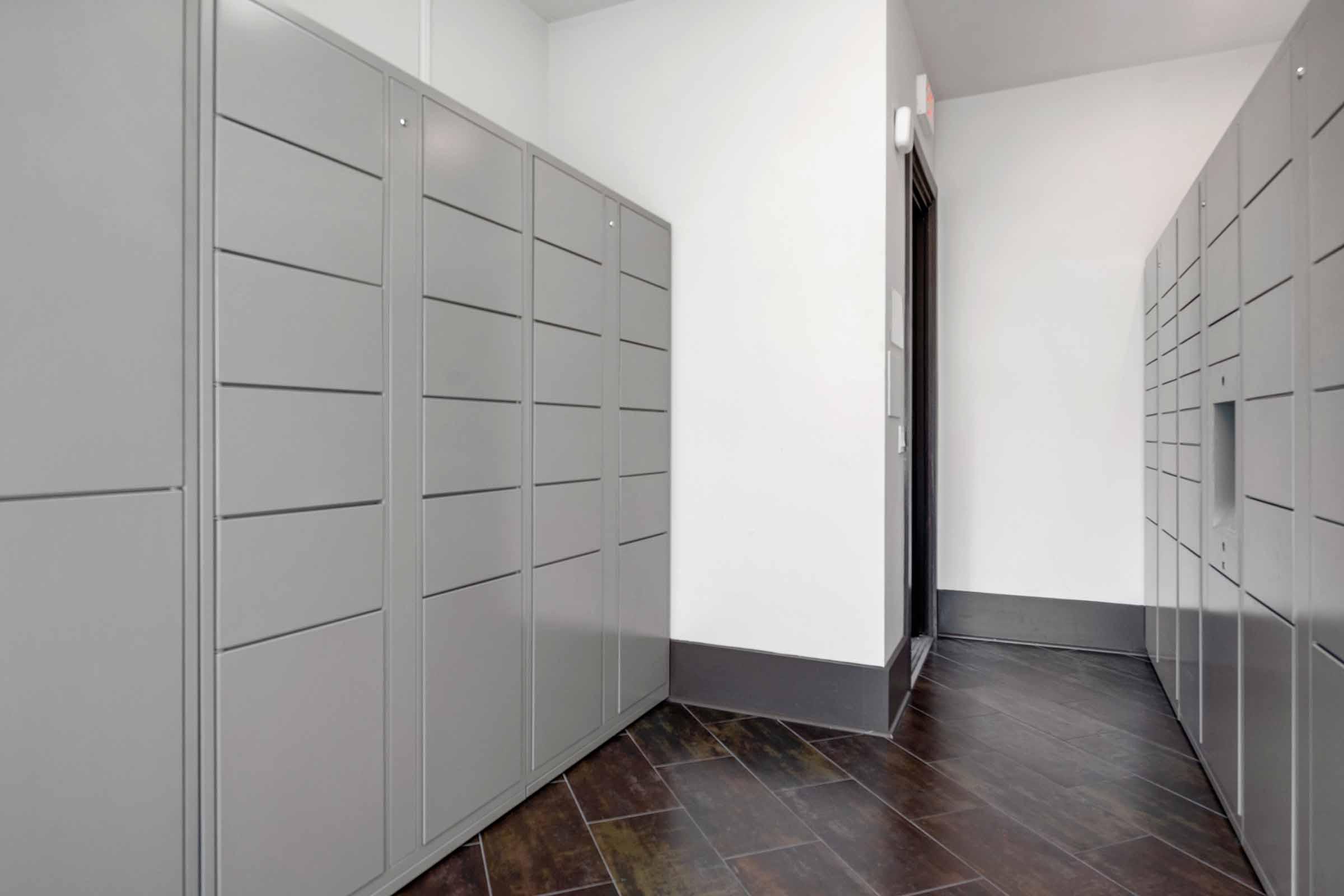 A corridor featuring gray lockers against a white wall, with a brown tiled floor. The lockers are metallic and organized in rows, providing storage solutions. Soft lighting enhances the modern and clean aesthetic of the space.