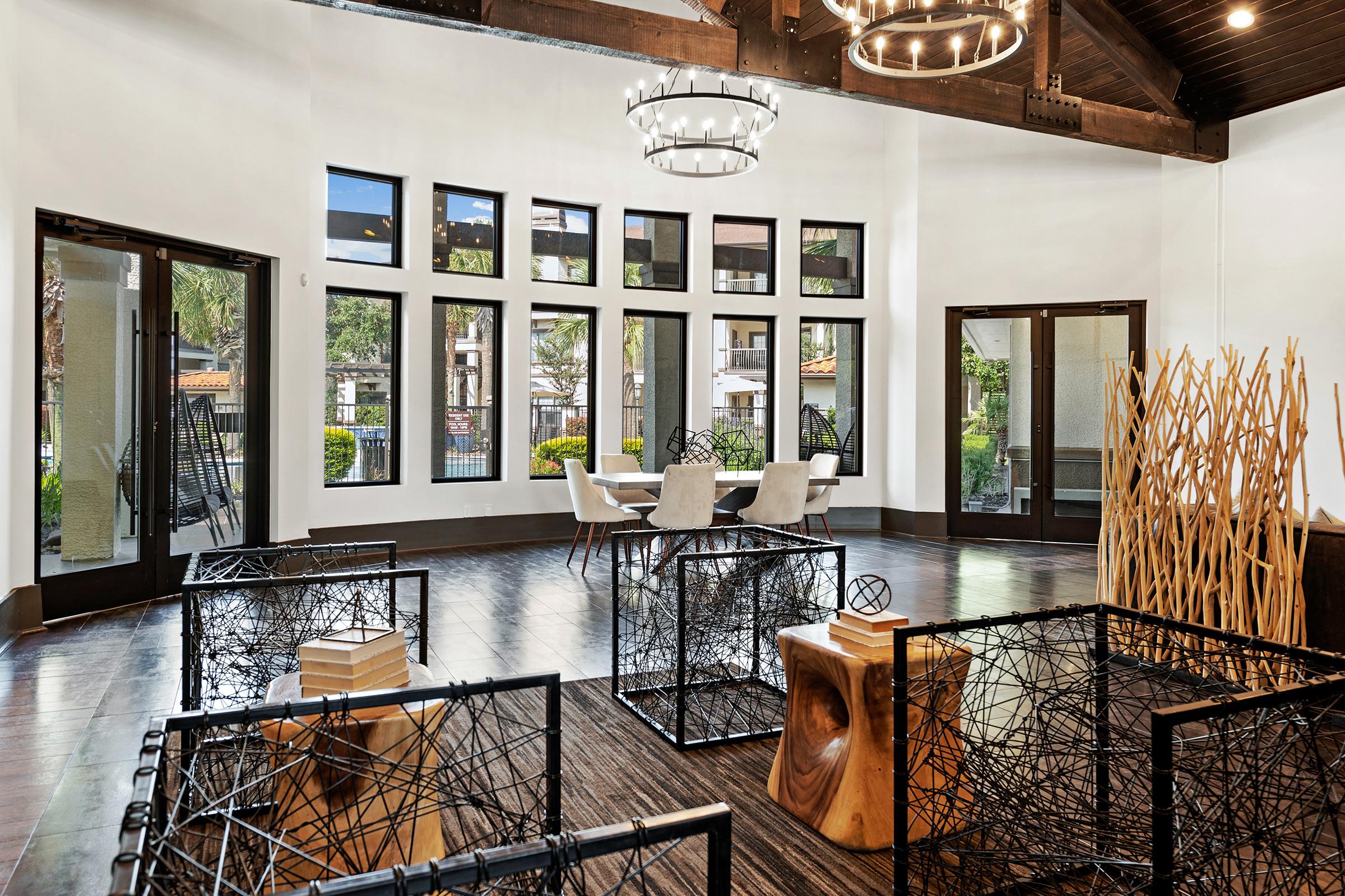 A modern, stylish interior of a lounge area featuring large windows, a wooden ceiling with a chandelier, and minimalist furniture. Black wire cube tables and a wooden stump serve as decorative elements, while a seating area with white chairs is visible in the background, enhancing the open, airy atmosphere.