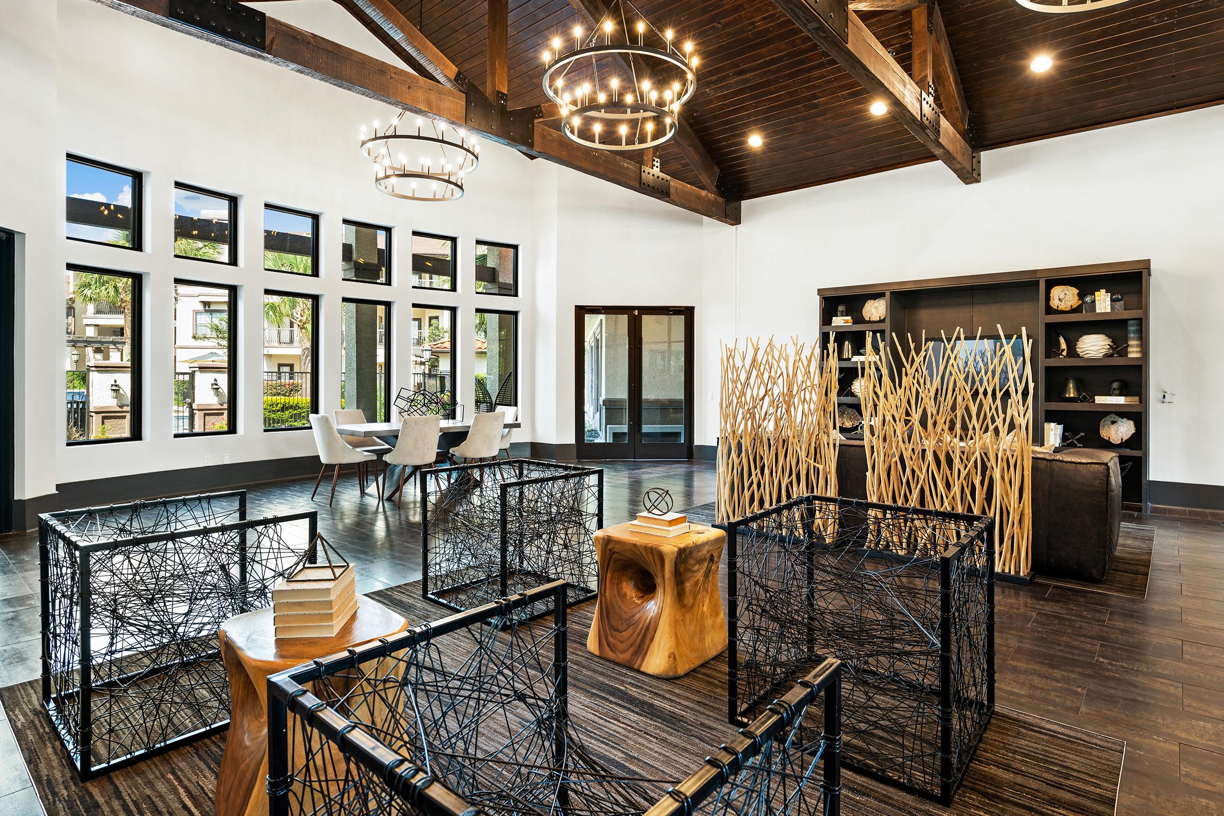 Modern interior of a spacious room featuring large windows, wooden beams, and decorative lighting fixtures. The furnishings include a dark wooden bookshelf, wooden stools, and open seating area with white chairs. Decorative elements include spatial dividers made of wood and string-like designs, creating an inviting atmosphere.