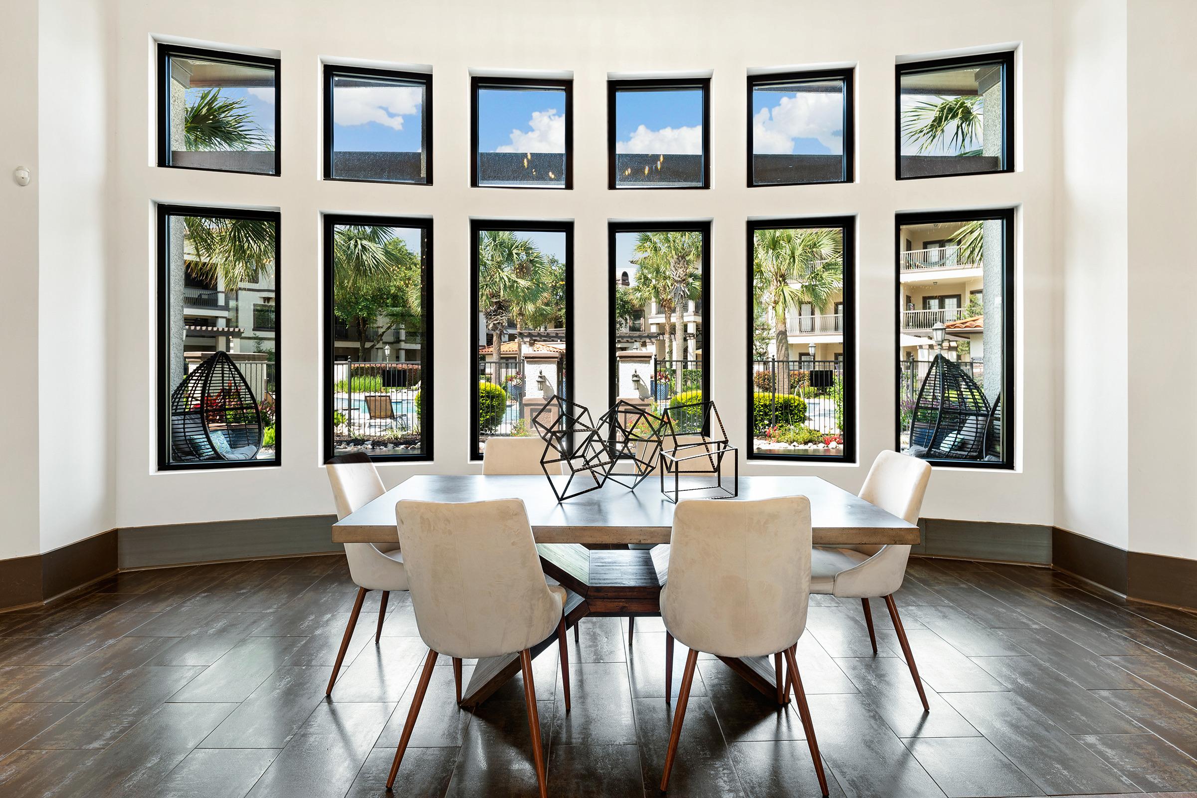 A modern dining area featuring a wooden table surrounded by four white chairs. Large windows allow natural light to flood the space, showcasing views of palm trees and an outdoor pool area. Stylish geometric decor is displayed on the table, adding an artistic touch to the inviting setting.