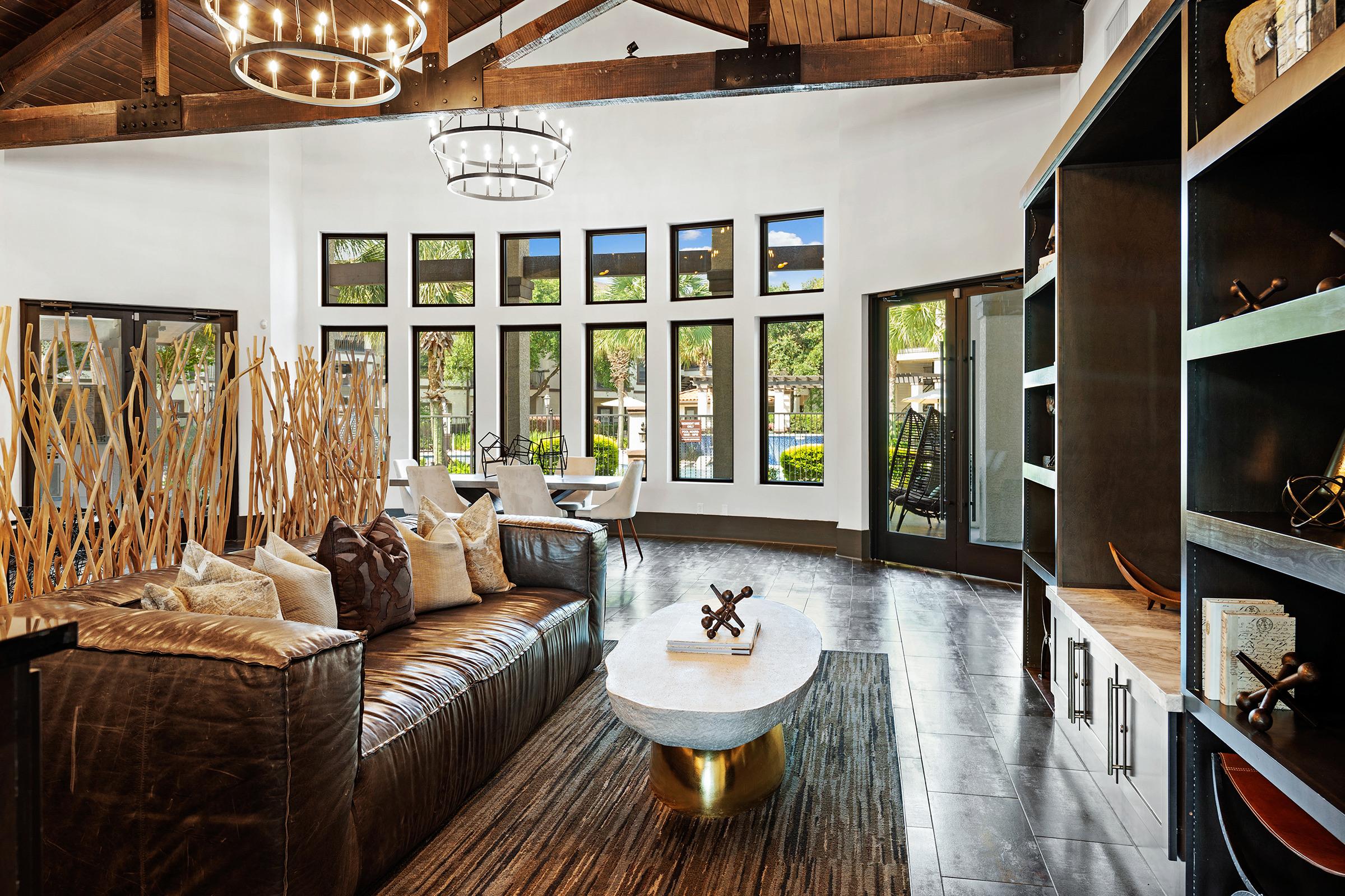 A modern living space featuring a brown leather sofa with decorative pillows, a stylish coffee table, and large windows that provide natural light. There are wooden beams on the ceiling, decorative shelving, and a dining area in the background with white chairs and a view of greenery outside.