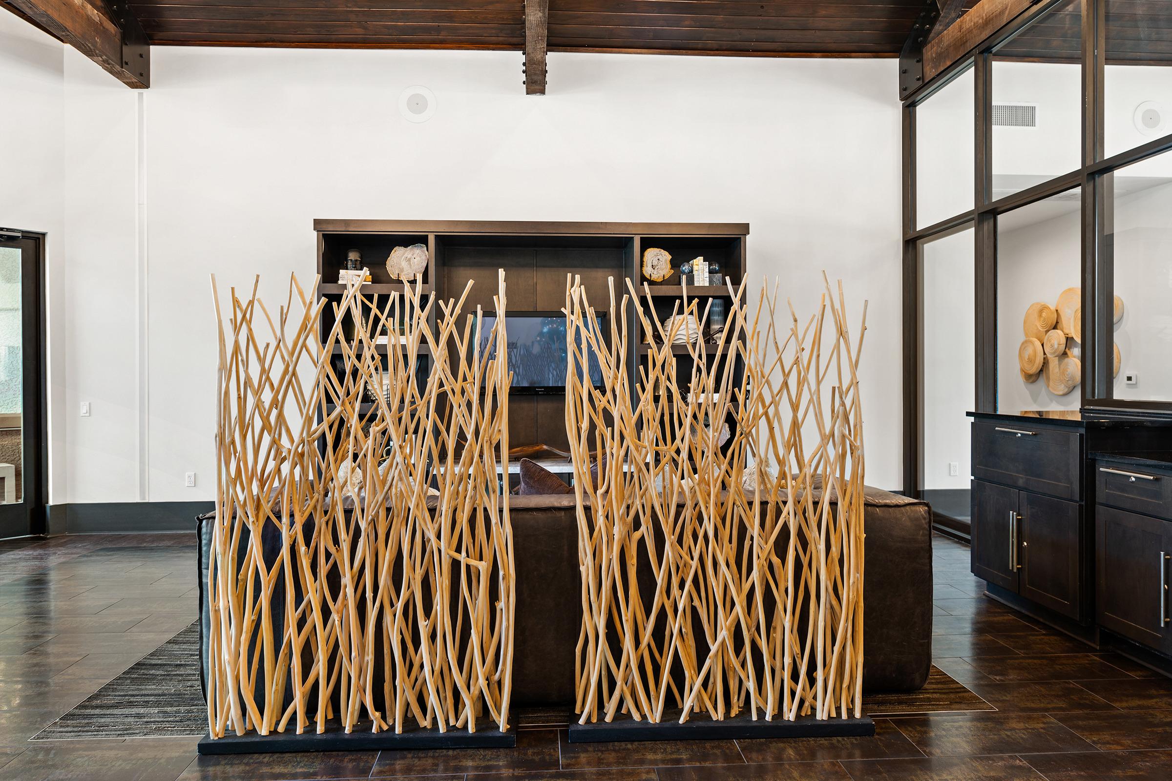 A modern interior space featuring a wooden decor, including tall brown stick-like sculptures arranged in two vertical clusters. The background showcases a dark wooden shelf with decorative items and a large flat-screen TV. The room has high ceilings with exposed beams and a sleek, dark floor.