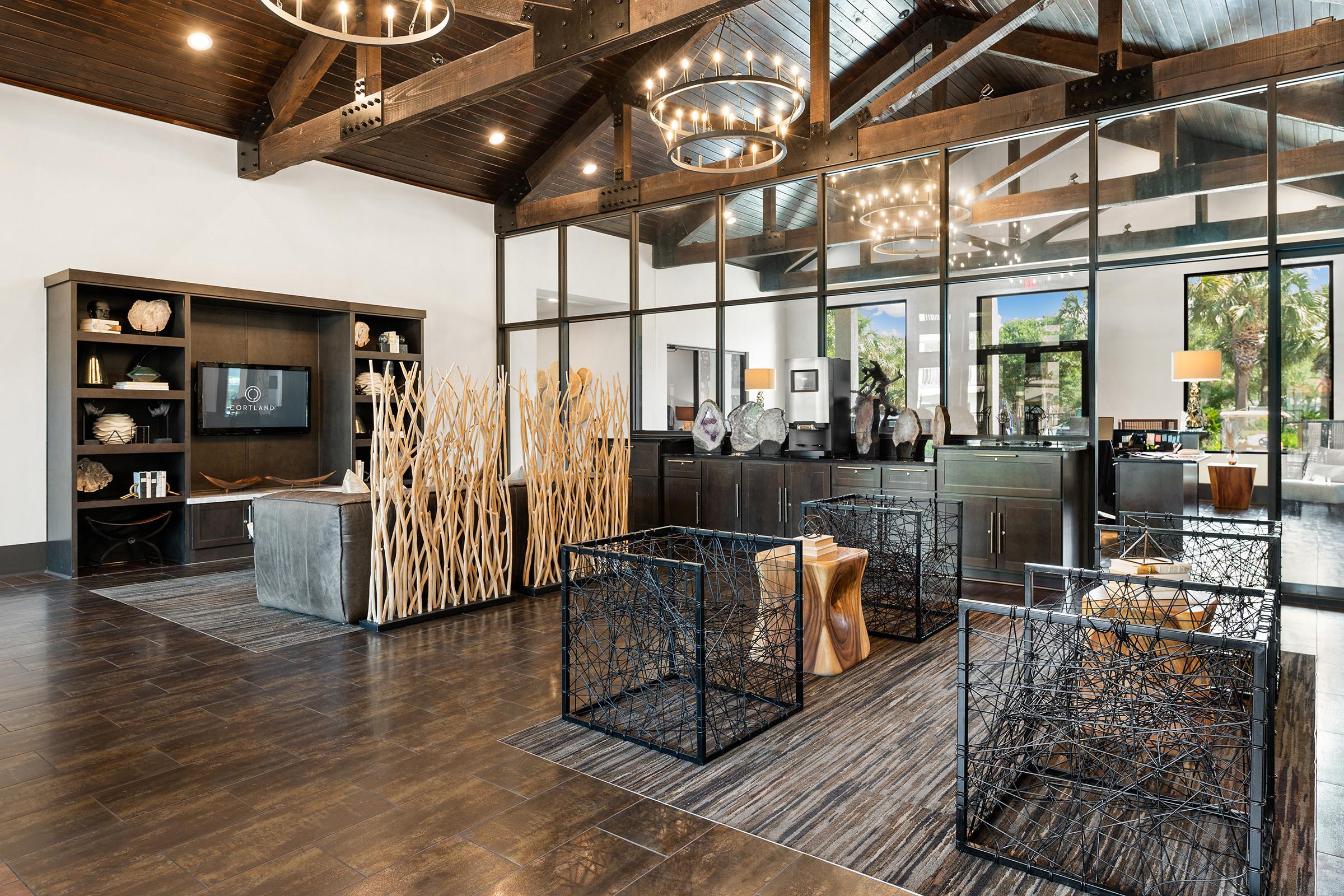 Modern interior of a stylish lobby featuring wooden beams, large glass panels, and contemporary furniture. Decorative elements include woven centerpieces and elegant light fixtures. The space is bright and welcoming, with artwork displayed on shelves and a comfortable seating area.