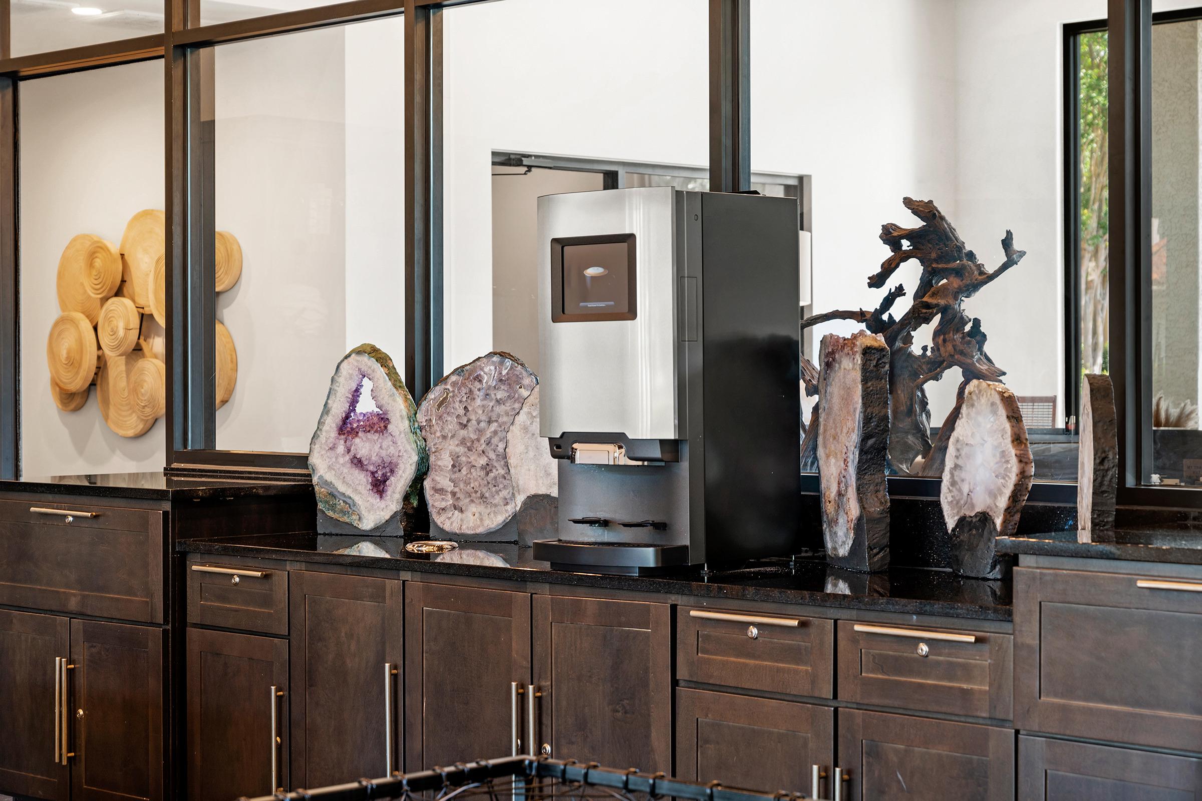 A stylish kitchen area featuring a modern appliance, surrounded by large, decorative geodes. The geodes exhibit various shades of purple and clear crystals, adding an elegant touch to the dark cabinetry and counter. The background includes a set of wooden wall decor, enhancing the contemporary design.