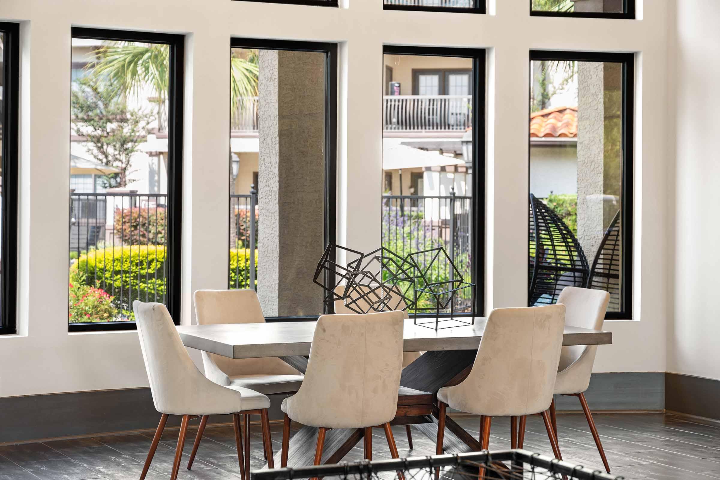 A modern dining area featuring a rectangular wooden table surrounded by six upholstered chairs. Large windows offer a view of greenery outside. A geometric sculpture is positioned on the table, adding an artistic touch to the space. The decor is bright and contemporary, enhancing the inviting atmosphere.