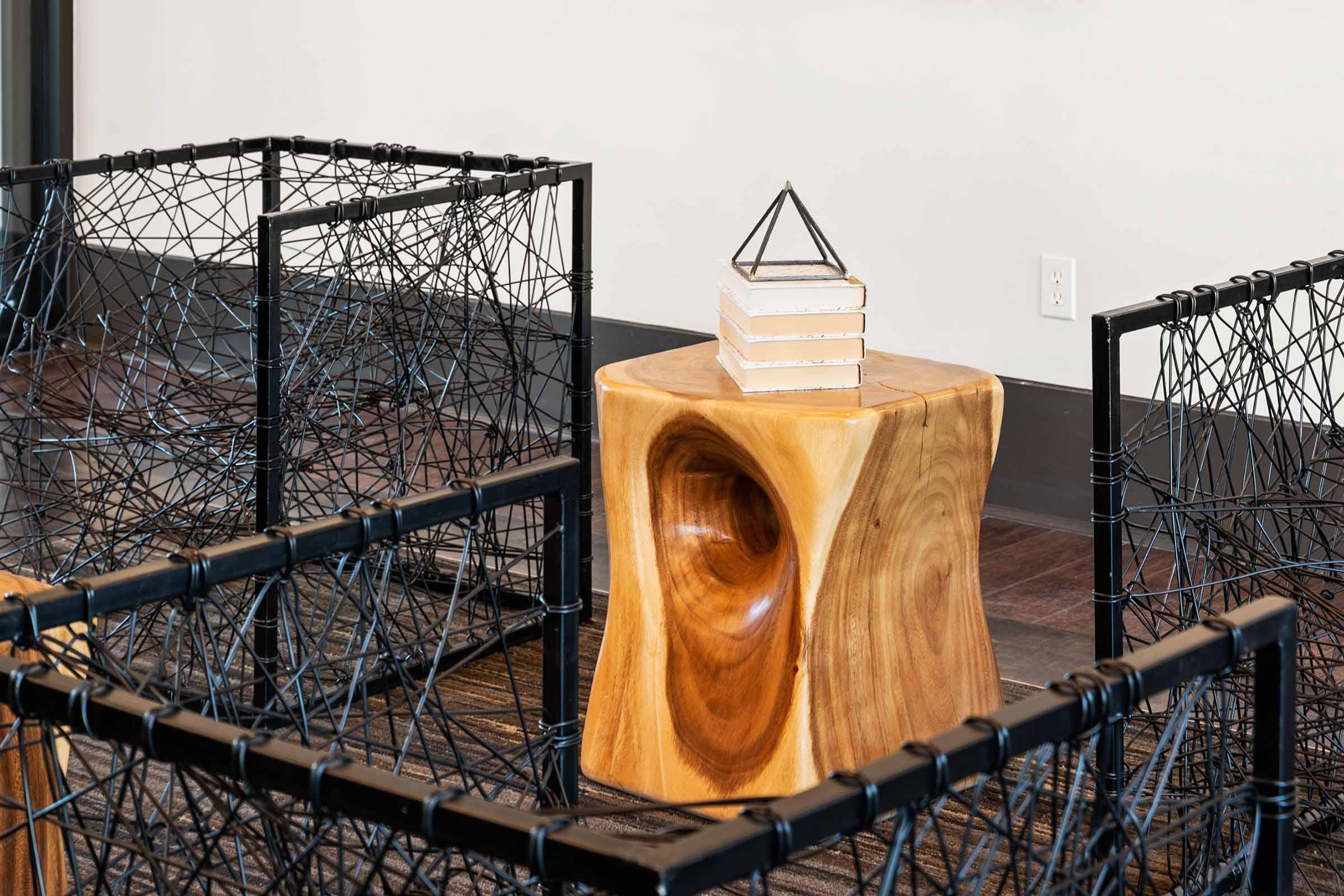 A wooden stool with a hollowed center stands next to a small stack of white books topped with a geometric sculpture. The background features black wire structures forming a geometric enclosure, creating a modern and artistic interior setting.