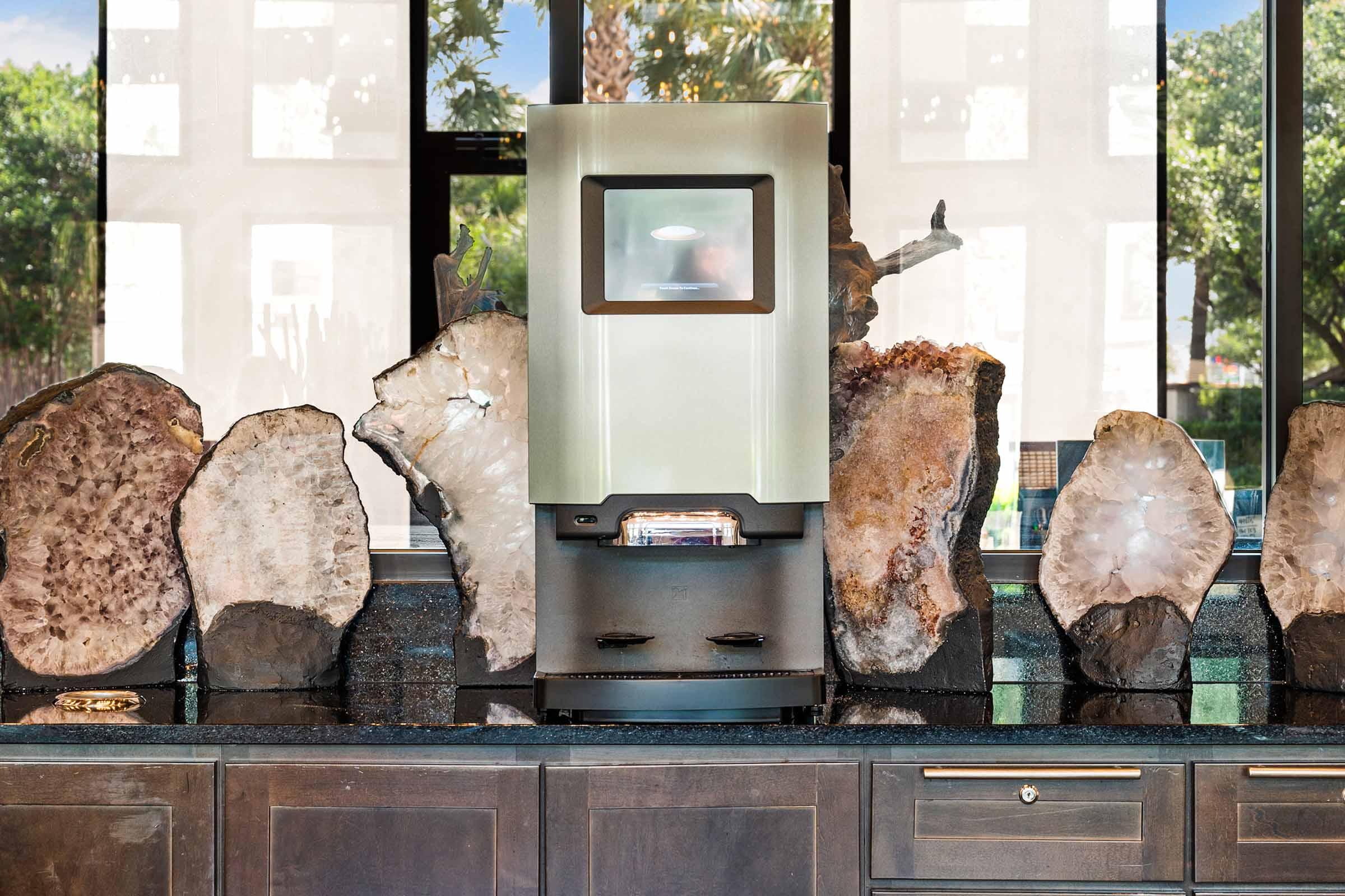 A modern ice maker is stationed on a dark countertop, flanked by large, polished stone slabs. The background features large windows with greenery visible outside, adding a bright, natural ambiance to the setting. The overall design combines contemporary functionality with natural materials.