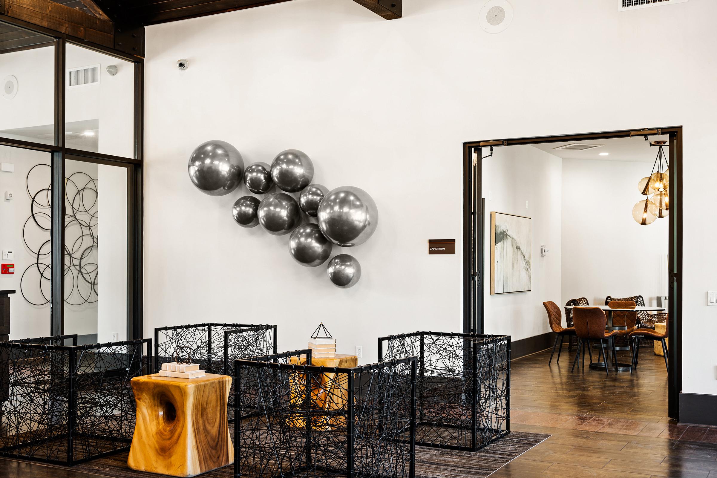 Interior of a modern space featuring a wall with a cluster of metallic gray balloons. There are black wire-frame furniture pieces arranged in the foreground, and a doorway leads to a dining area with chairs and a table visible. The walls are light-colored, enhancing the contemporary feel of the design.