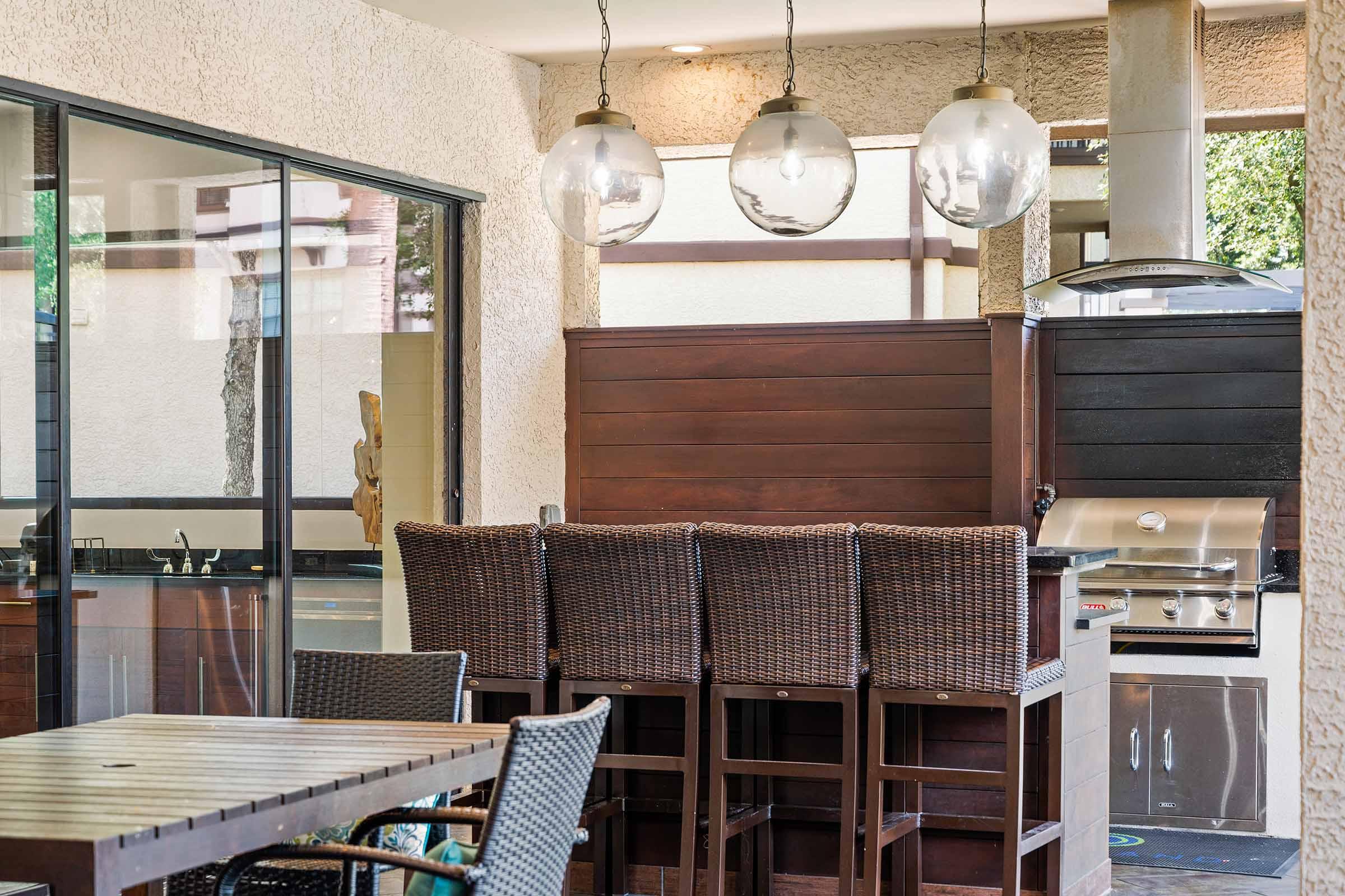 An outdoor kitchen area featuring modern bar stools, a wooden dining table, and a built-in grill. Large glass lights hang above the bar, and there's a glass wall providing a view of the surroundings. The space has a warm, inviting ambiance, suitable for entertaining.