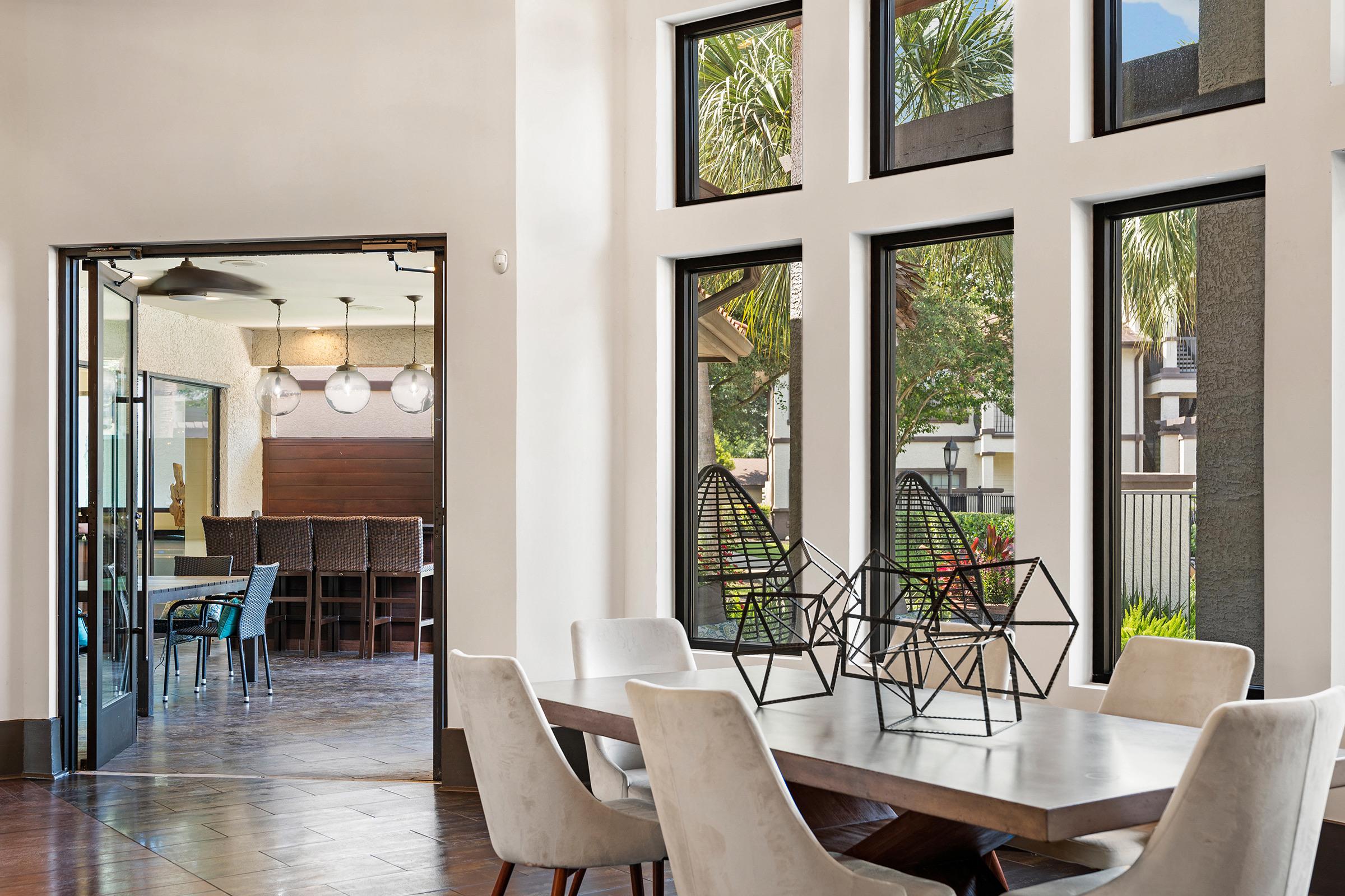 A bright, modern dining area featuring a large wooden table surrounded by upholstered chairs. Geometric decor is displayed on the table. Large windows allow natural light to fill the space, with views of greenery outside. An open doorway leads to another room with a dining setup.