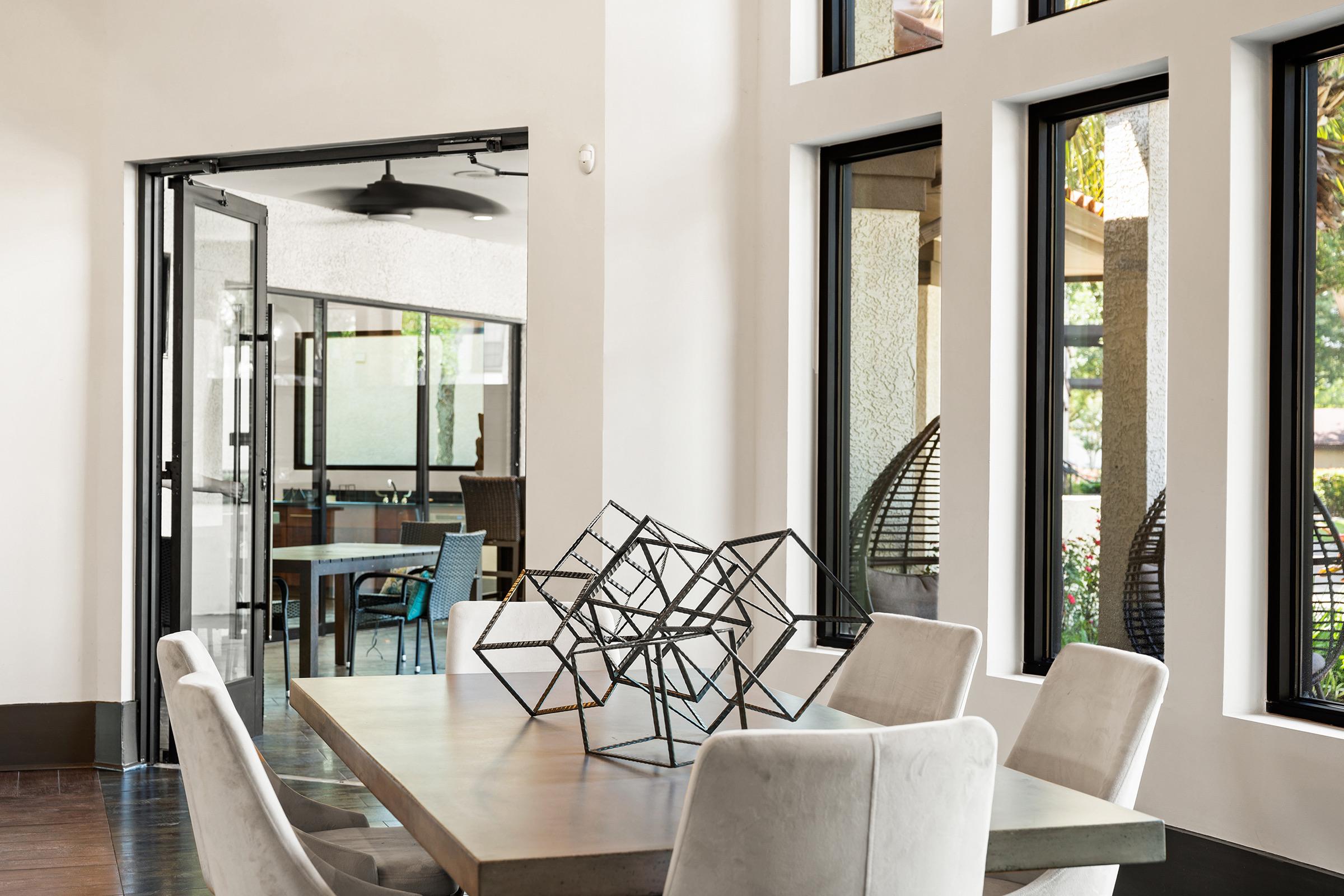 A modern dining area with a large rectangular table surrounded by upholstered chairs. A geometric metal sculpture sits at the center of the table. Large windows provide ample natural light, and the space opens to another room with additional seating and greenery visible outside.