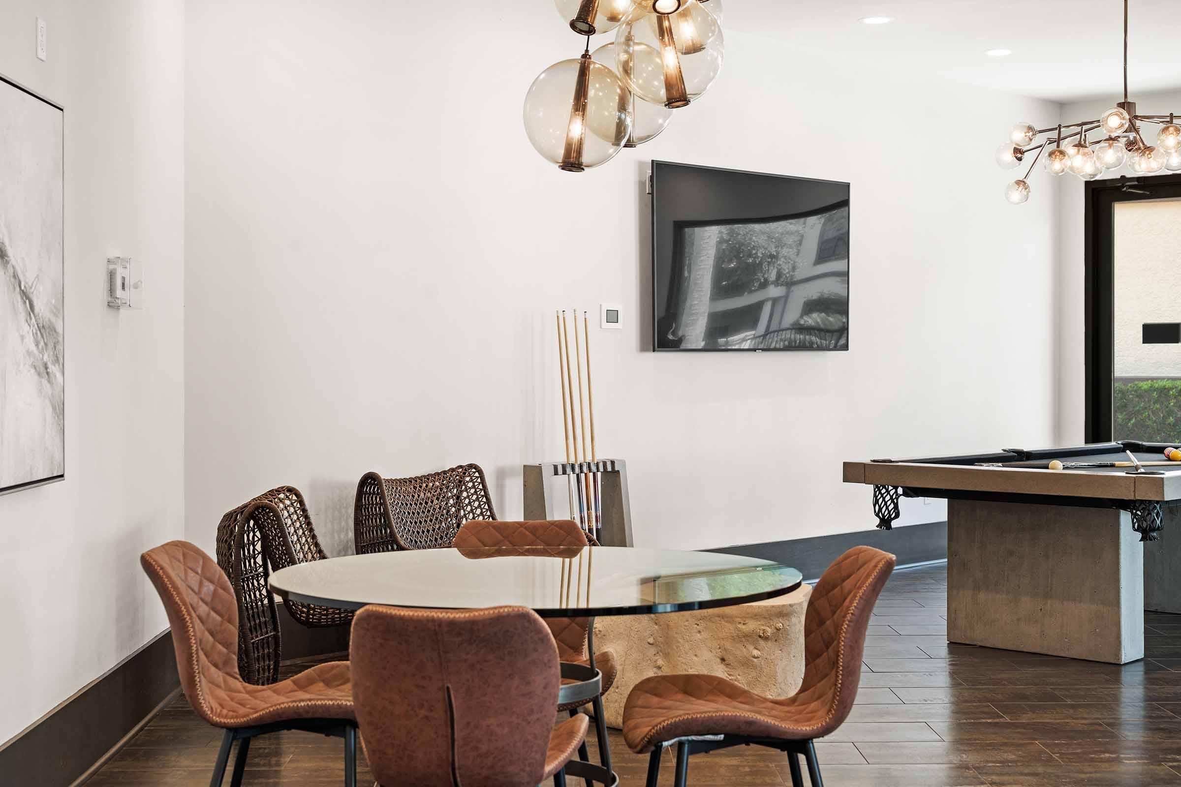 A modern dining area featuring a round glass table surrounded by stylish woven and upholstered chairs. In the background, there's a wall-mounted TV and a billiards table. The room has a bright, contemporary ambiance with sleek lighting fixtures and wooden flooring.