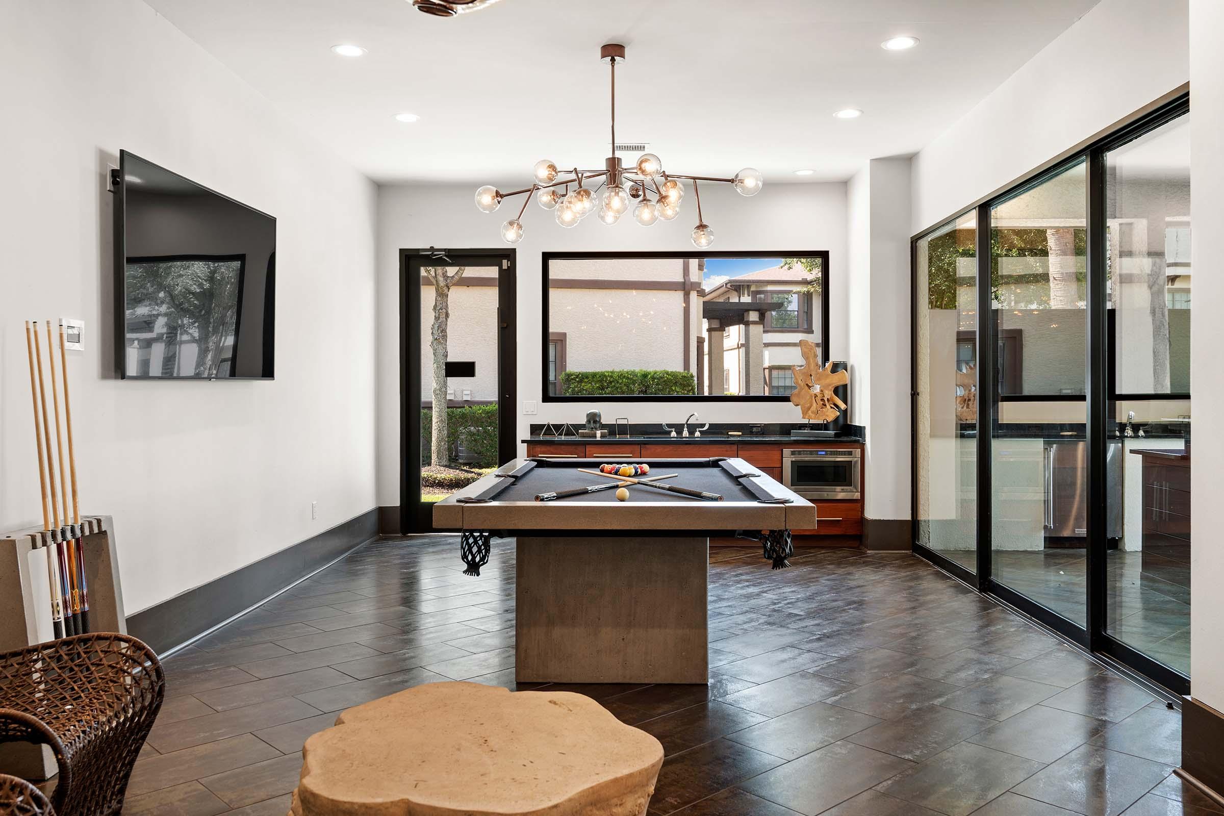 Bright and modern game room featuring a billiard table in the center, with a chandelier overhead and large windows along one wall. A television is mounted on the wall, and the space includes a kitchenette area. Natural light floods the room, highlighting the sleek design and contemporary furnishings.